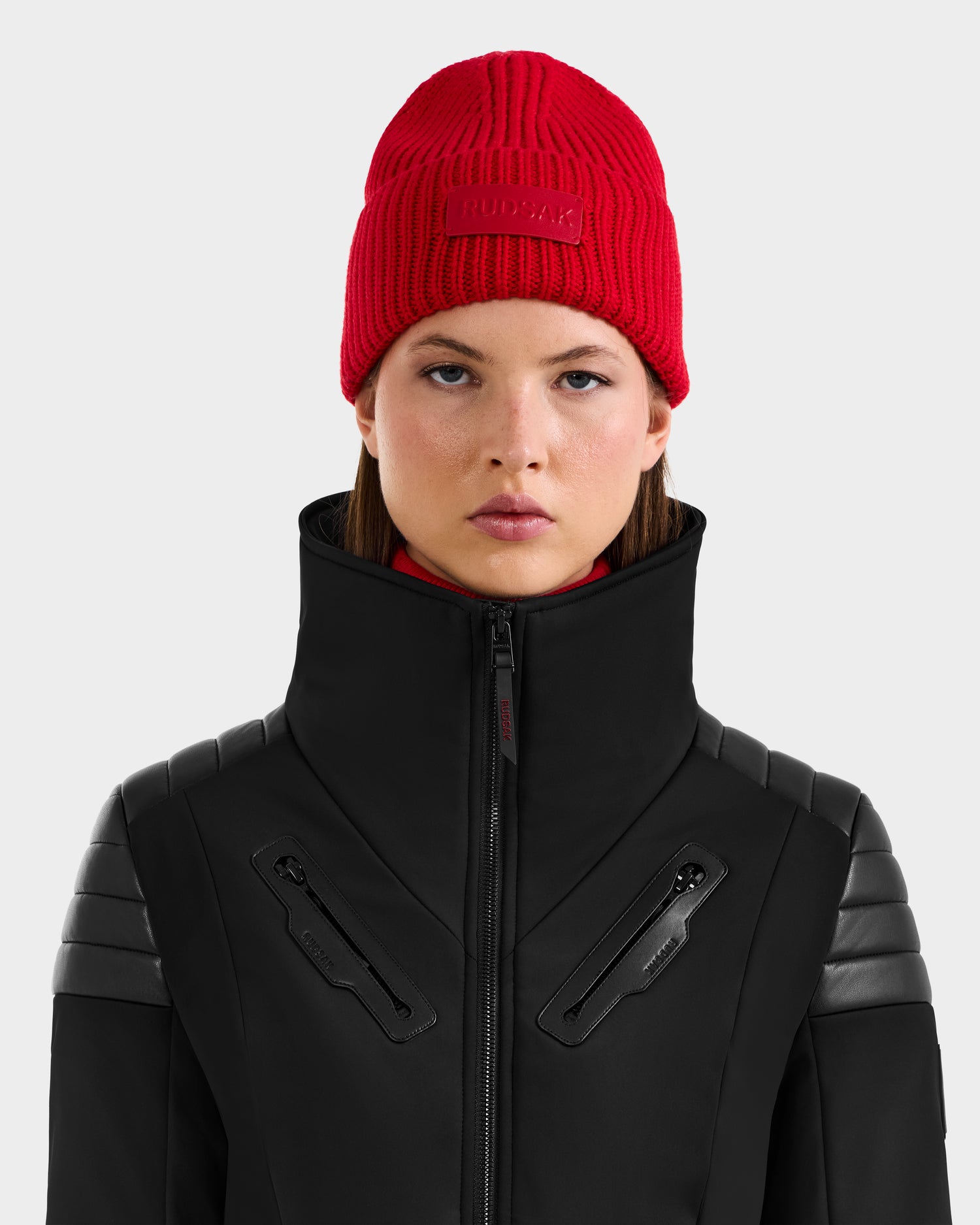 A person with straight brown hair wears the Rudsak COLIN - 9424518 Flame Red beanie and a black high-collar jacket with zippers, facing forward against a plain light gray background.