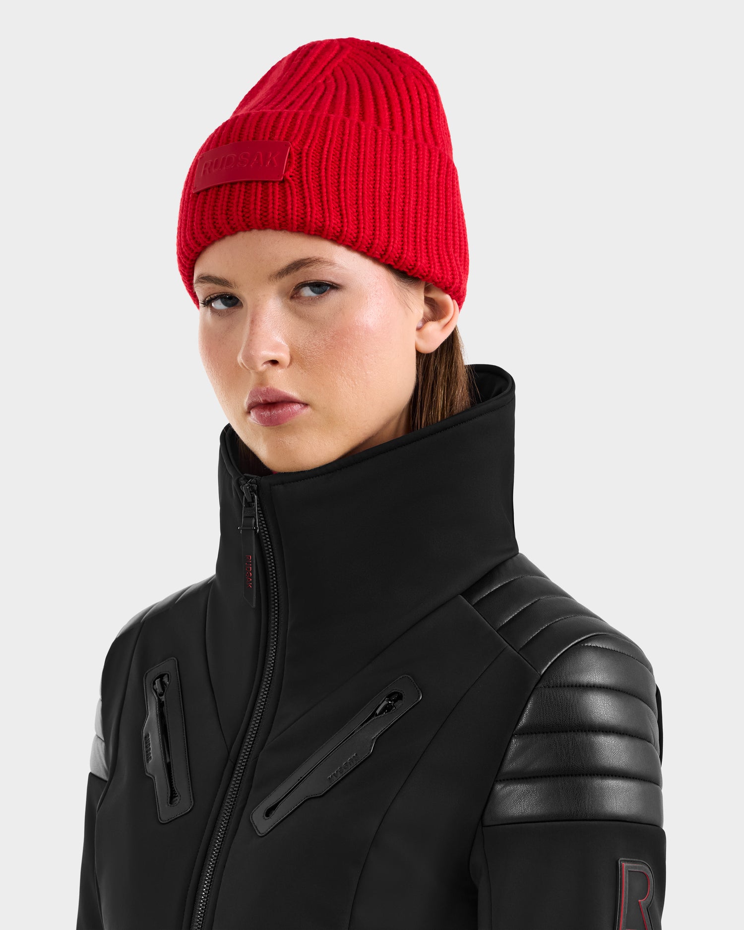 A woman wears the flame red Rudsak COLIN beanie and a black jacket with leather accents and a high collar, looking slightly to the side with a neutral expression against a plain background.