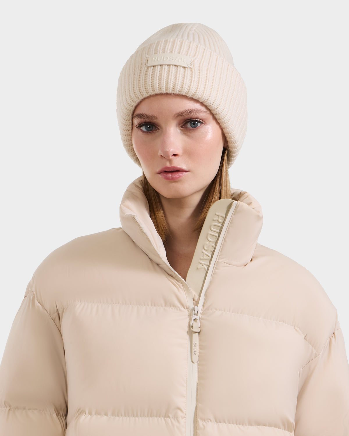 A woman wears the Rudsak COLIN - 9424518 Vanilla knit beanie, featuring a rectangular front label, paired with a beige high-collar puffer jacket against a plain white background.