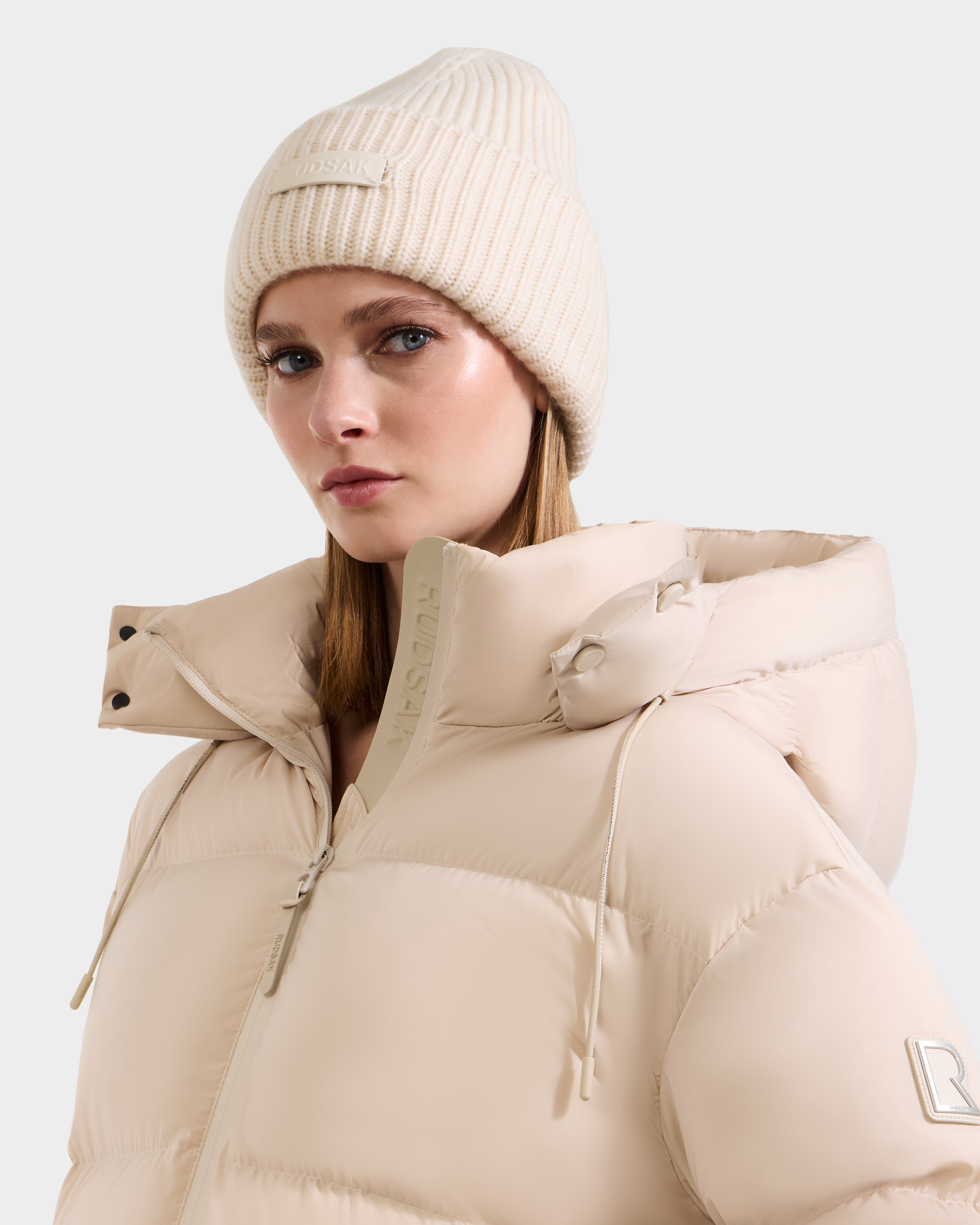 A woman in the Rudsak COLIN - 9424518 Vanilla puffer jacket with a high collar and a matching merino wool beanie looks to the side against a plain light background.