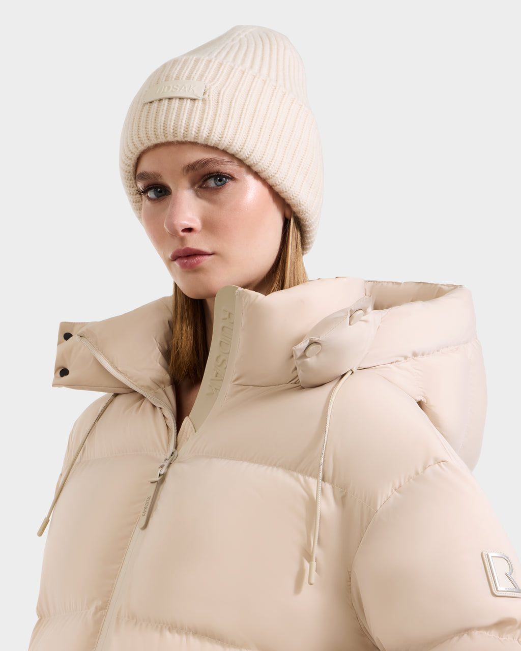 A woman in the Rudsak COLIN - 9424518 Vanilla puffer jacket with a high collar and a matching merino wool beanie looks to the side against a plain light background.