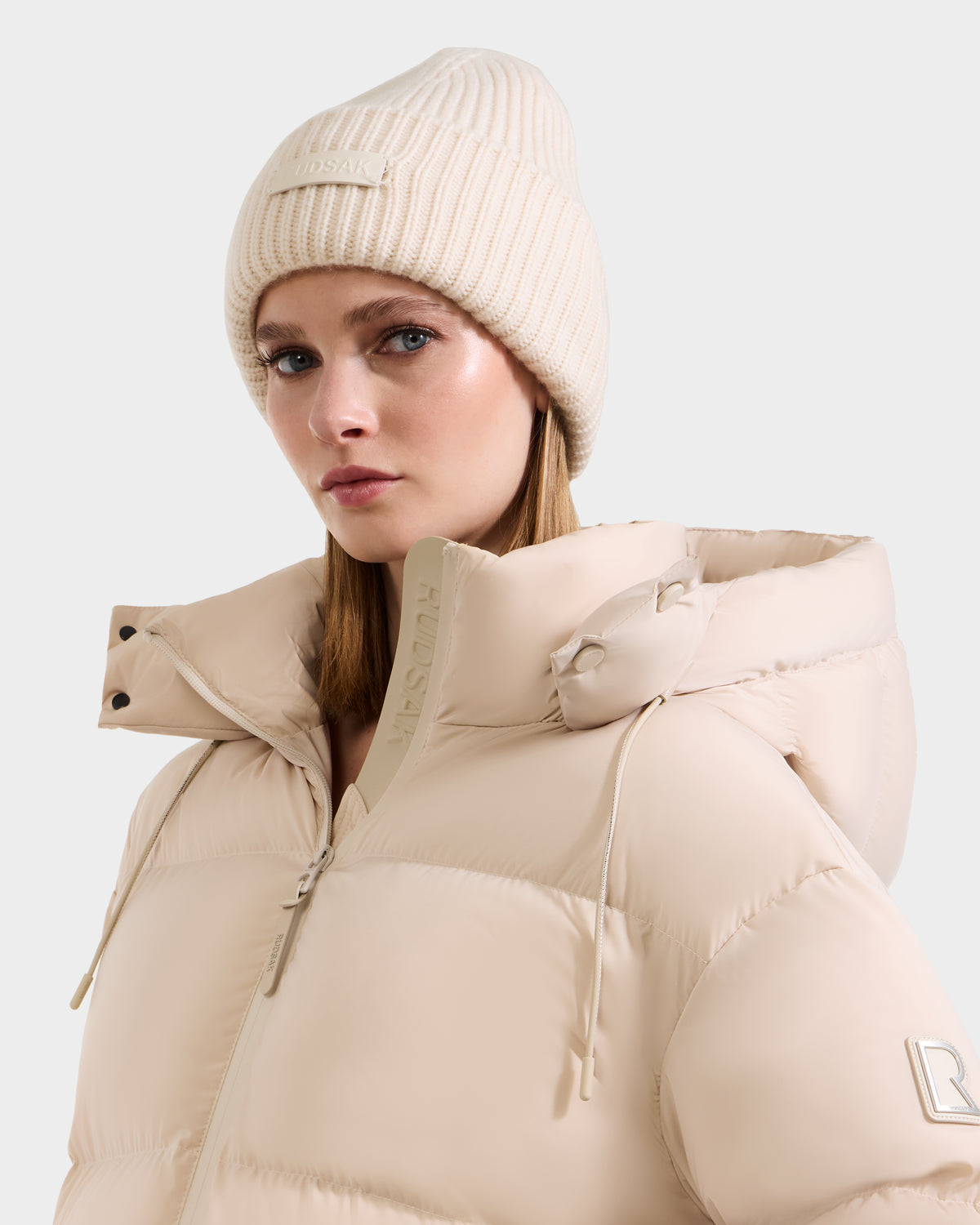 A woman in the Rudsak COLIN - 9424518 Vanilla puffer jacket with a high collar and a matching merino wool beanie looks to the side against a plain light background.