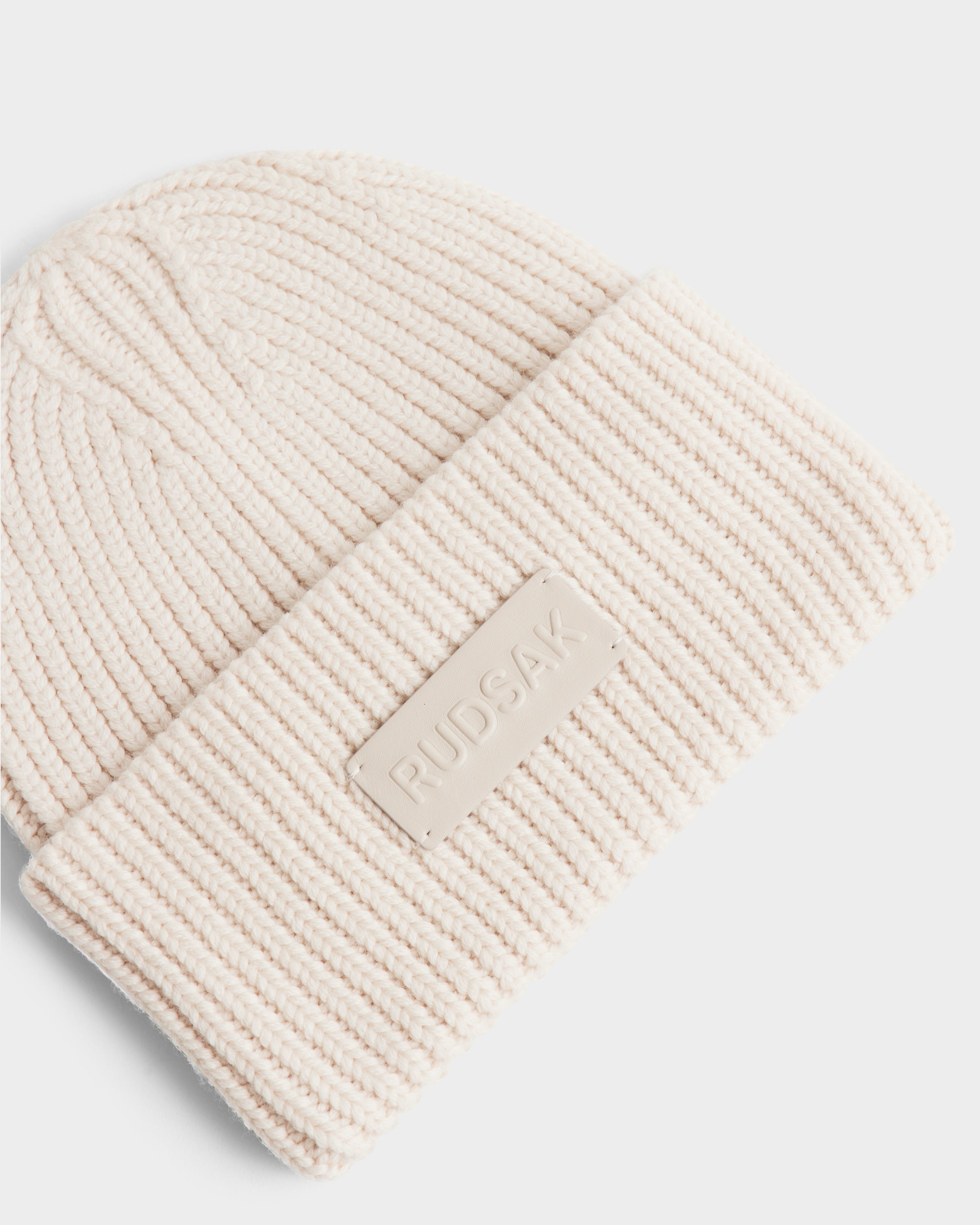 A close-up of the Rudsak COLIN - 9424518 Vanilla, a cream-colored unisex ribbed knit beanie with a folded brim and a rectangular Rudsak patch.