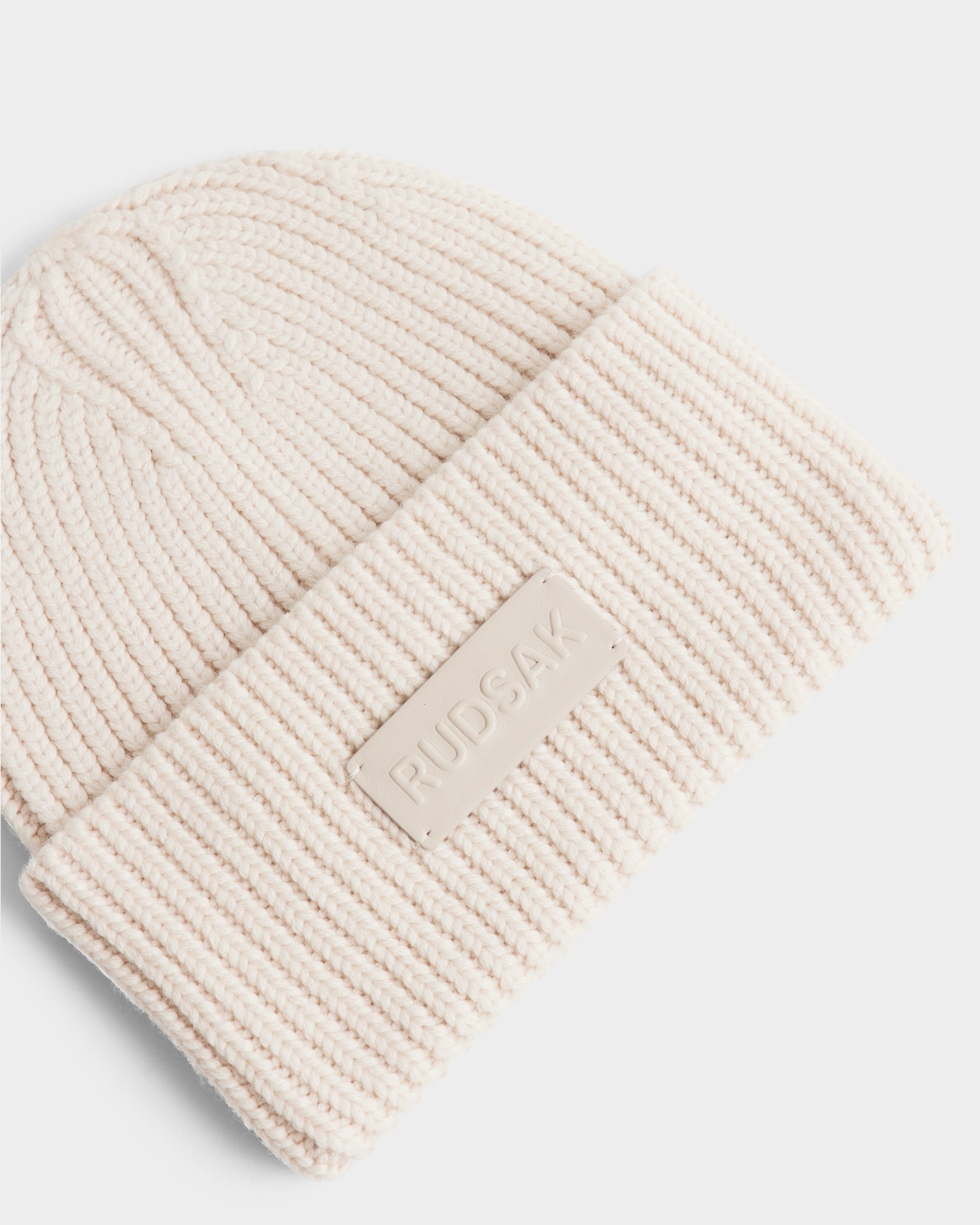 A close-up of the Rudsak COLIN - 9424518 Vanilla, a cream-colored unisex ribbed knit beanie with a folded brim and a rectangular Rudsak patch.