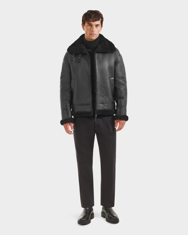 A man faces forward in a black Rudsak CONOR - 6124596 shearling leather coat, paired with a black turtleneck, pants, and shoes against a white background—an essential winter wardrobe look.