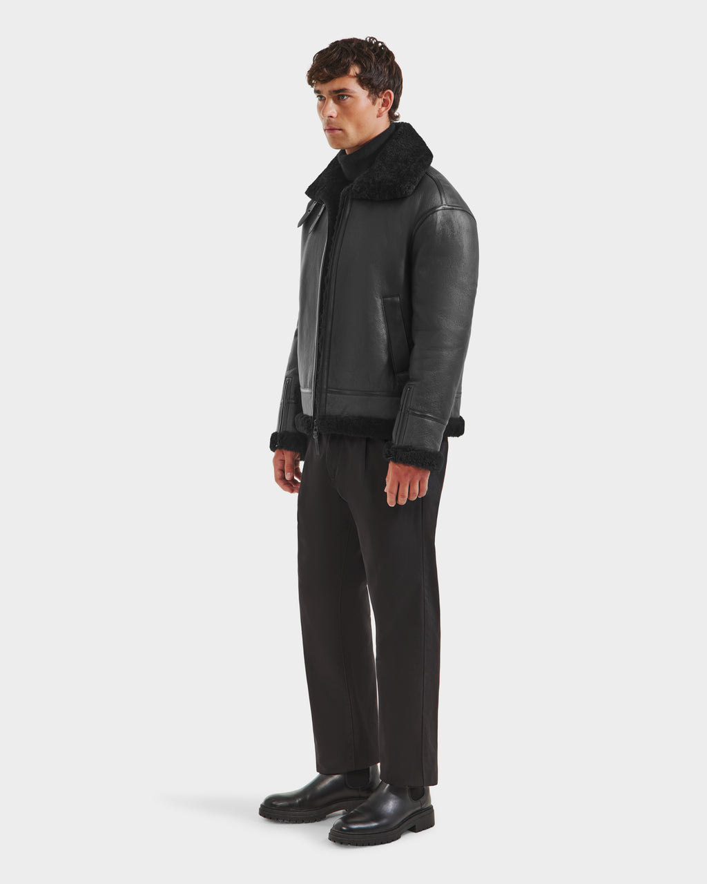 A man stands against a plain white background wearing the Rudsak CONOR - 6124596 Black mens shearling leather coat with a shearling collar, paired with black trousers and shoes—embodying a classic winter wardrobe staple.