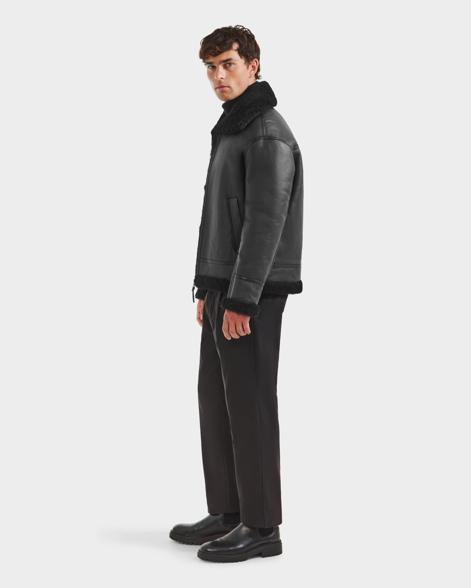 A man stands sideways wearing the Rudsak CONOR - 6124596 Black men’s shearling leather coat, paired with black pants and boots, against a plain white background.