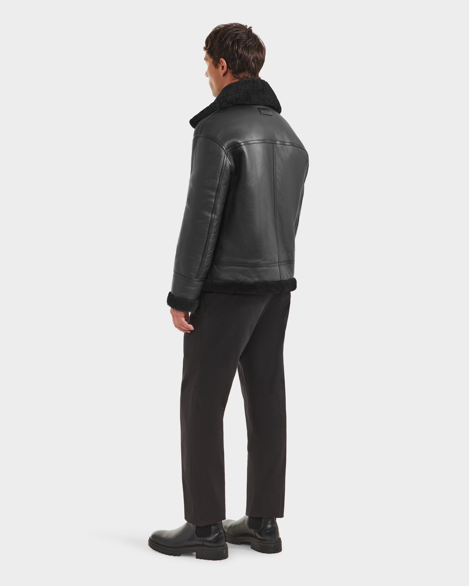 A man faces away, wearing the Rudsak CONOR - 6124596 Black shearling collar jacket with black pants and boots—a refined winter look—set against a plain white background.