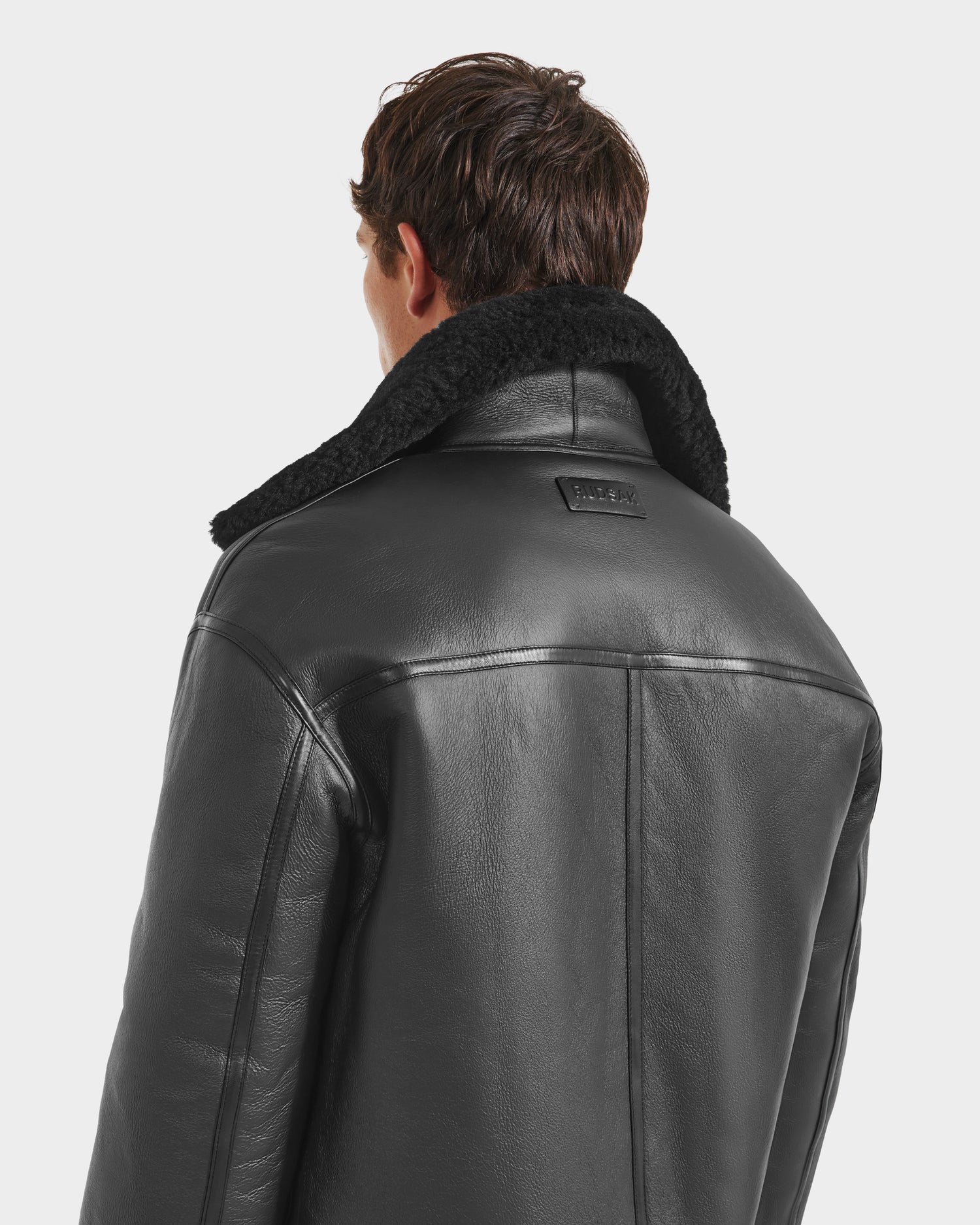 A person with short brown hair is seen from behind wearing the Rudsak CONOR - 6124596 Black men’s shearling leather coat with a textured finish and black shearling collar, perfect for upgrading any winter wardrobe. Plain white background.