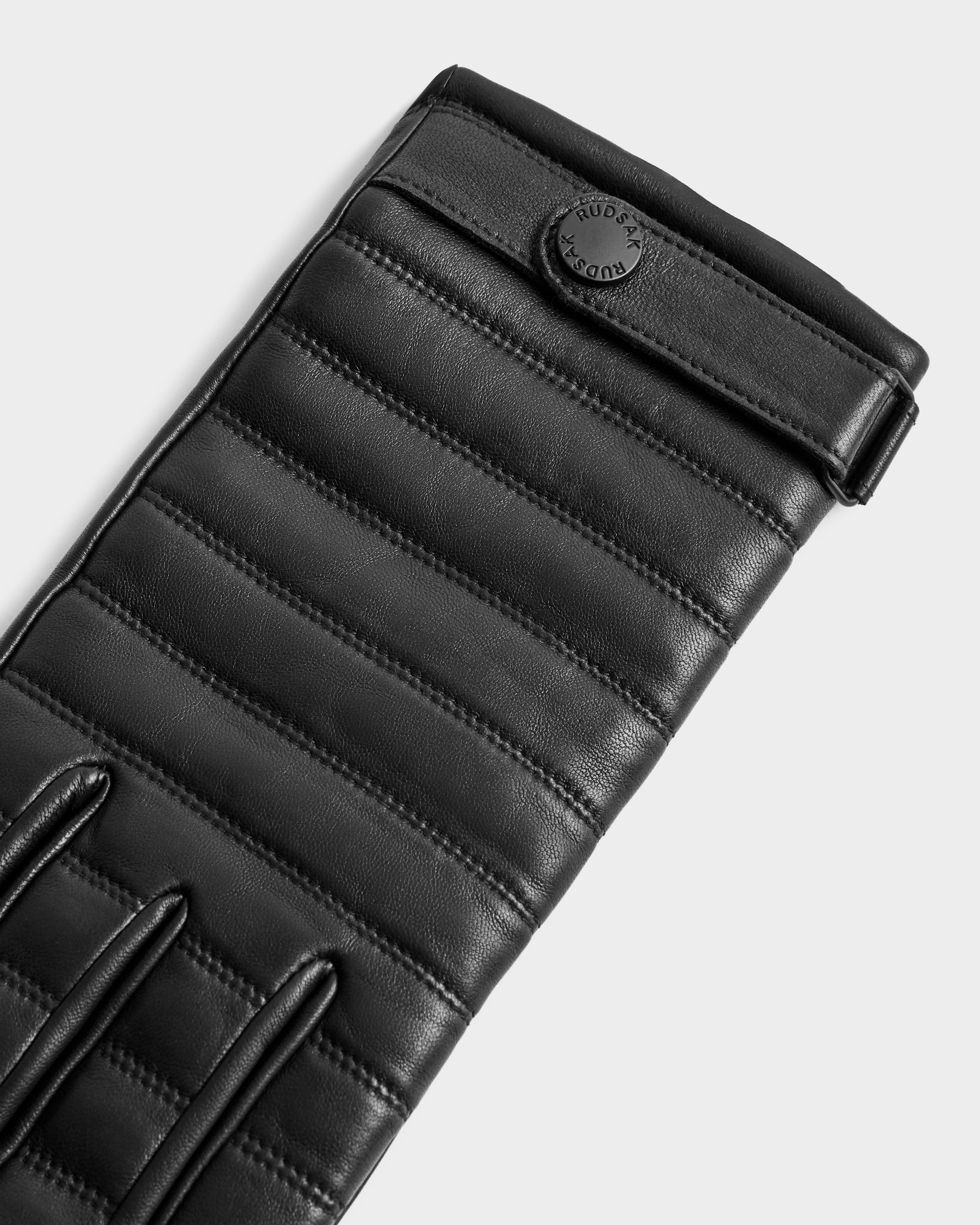 A close-up of the Rudsak CORTES - 6425036 Black quilted leather glove with horizontal stitching and a circular black button. These stylish leather gloves are perfect winter accessories against a plain, light-colored background.