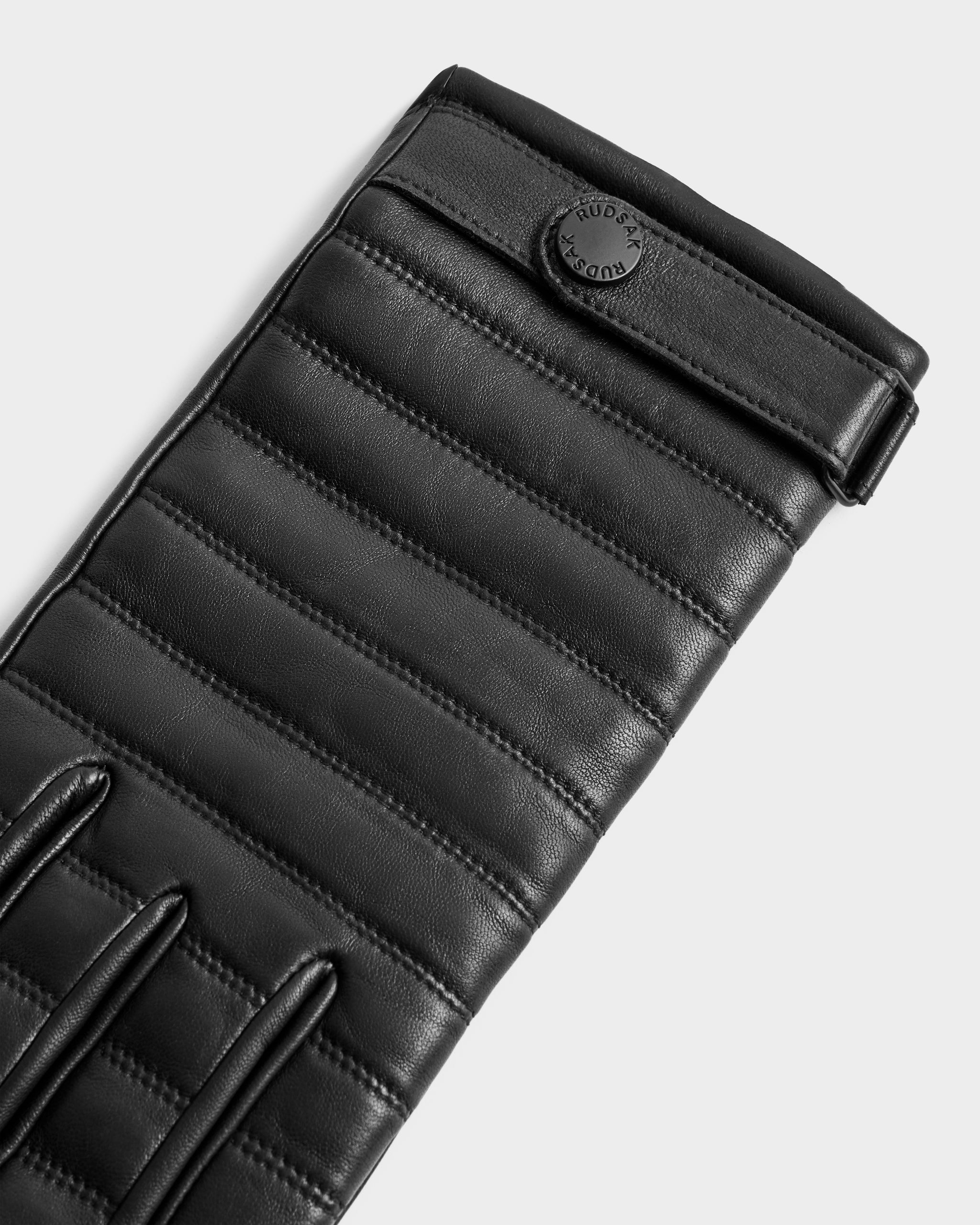 A close-up of the Rudsak CORTES - 6425036 Black quilted leather glove with horizontal stitching and a circular black button. These stylish leather gloves are perfect winter accessories against a plain, light-colored background.