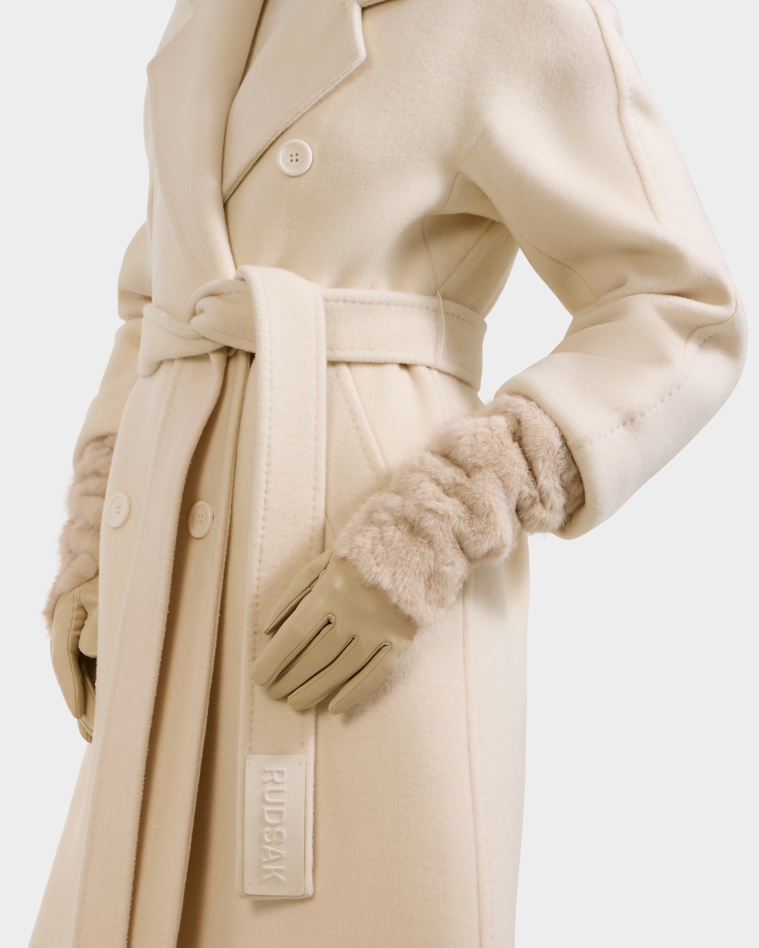 A person wearing the Rudsak COVE - 8425533 Vanilla cream wool coat with a belted waist, double-breasted buttons, faux fur cuffs, and beige leather gloves. "RUDSAK" is embossed on the belt. The person's face is not visible.
