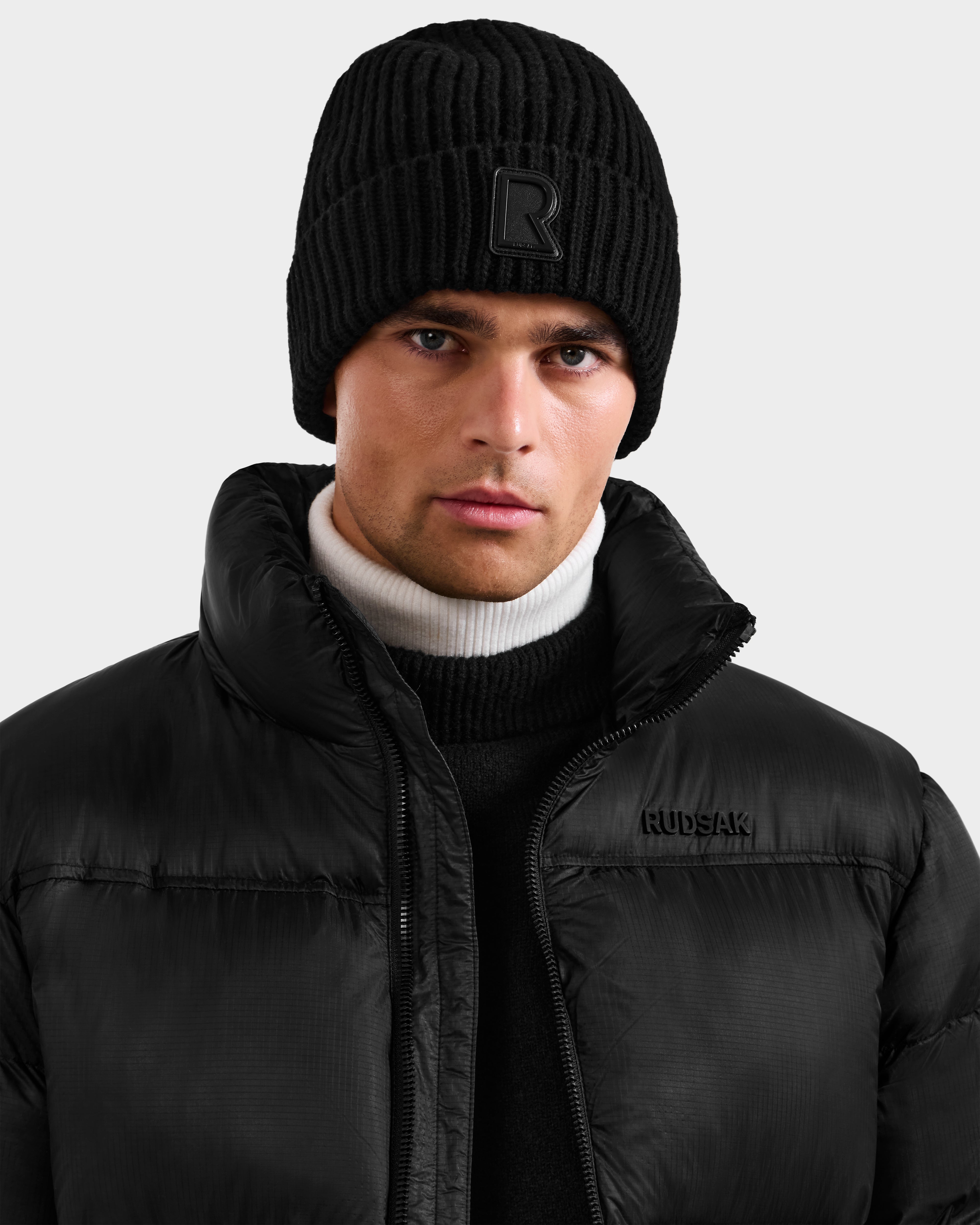A man wears the Rudsak CYBELLE - 9424504 Black knit beanie, paired with a Rudsak logo puffer jacket and a black and white turtleneck, standing against a plain light background to showcase this stylish unisex hat.