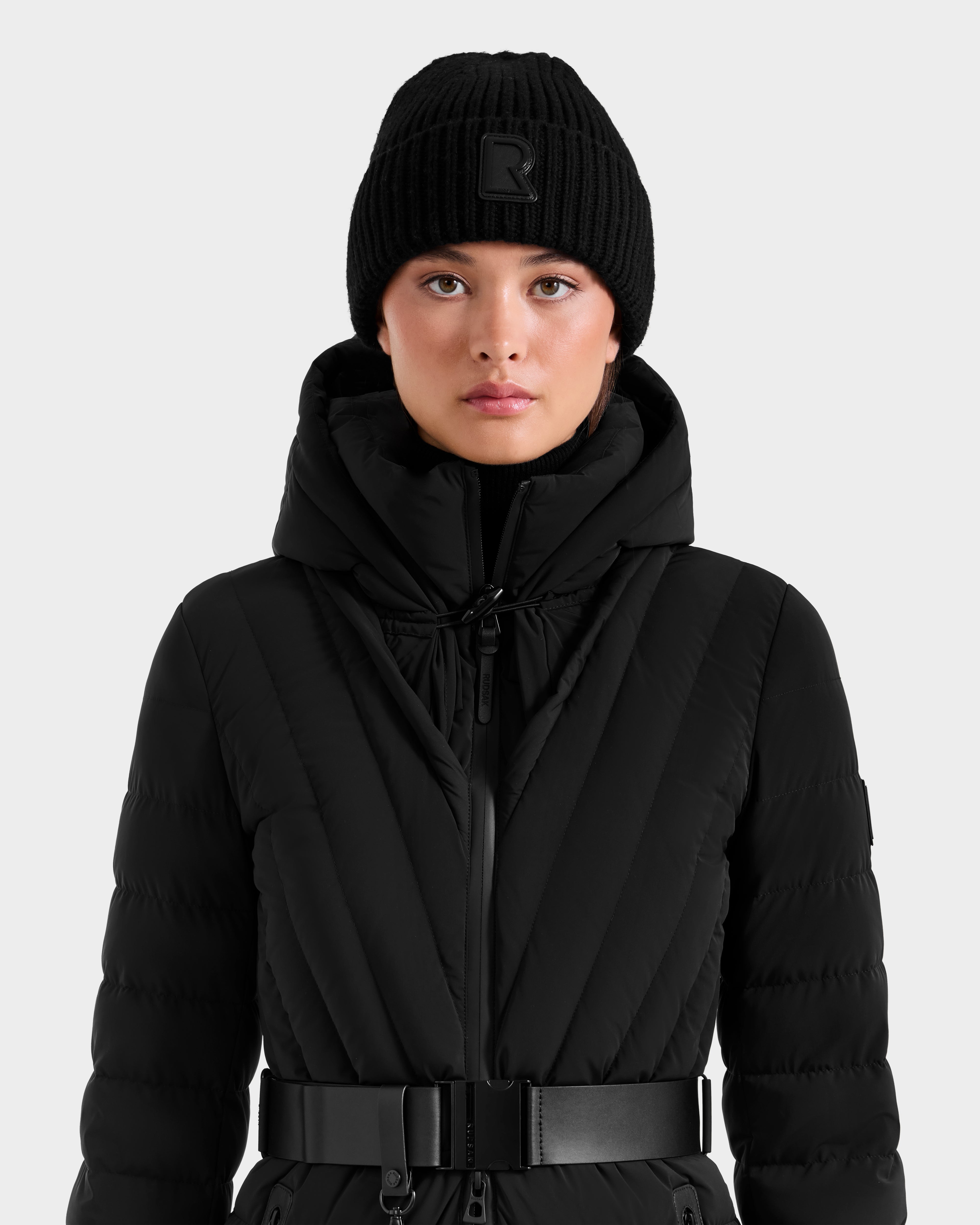 A woman wears the Rudsak CYBELLE - 9424504 Black quilted winter coat with a hood and a unisex knit hat, looking directly at the camera against a plain light background.