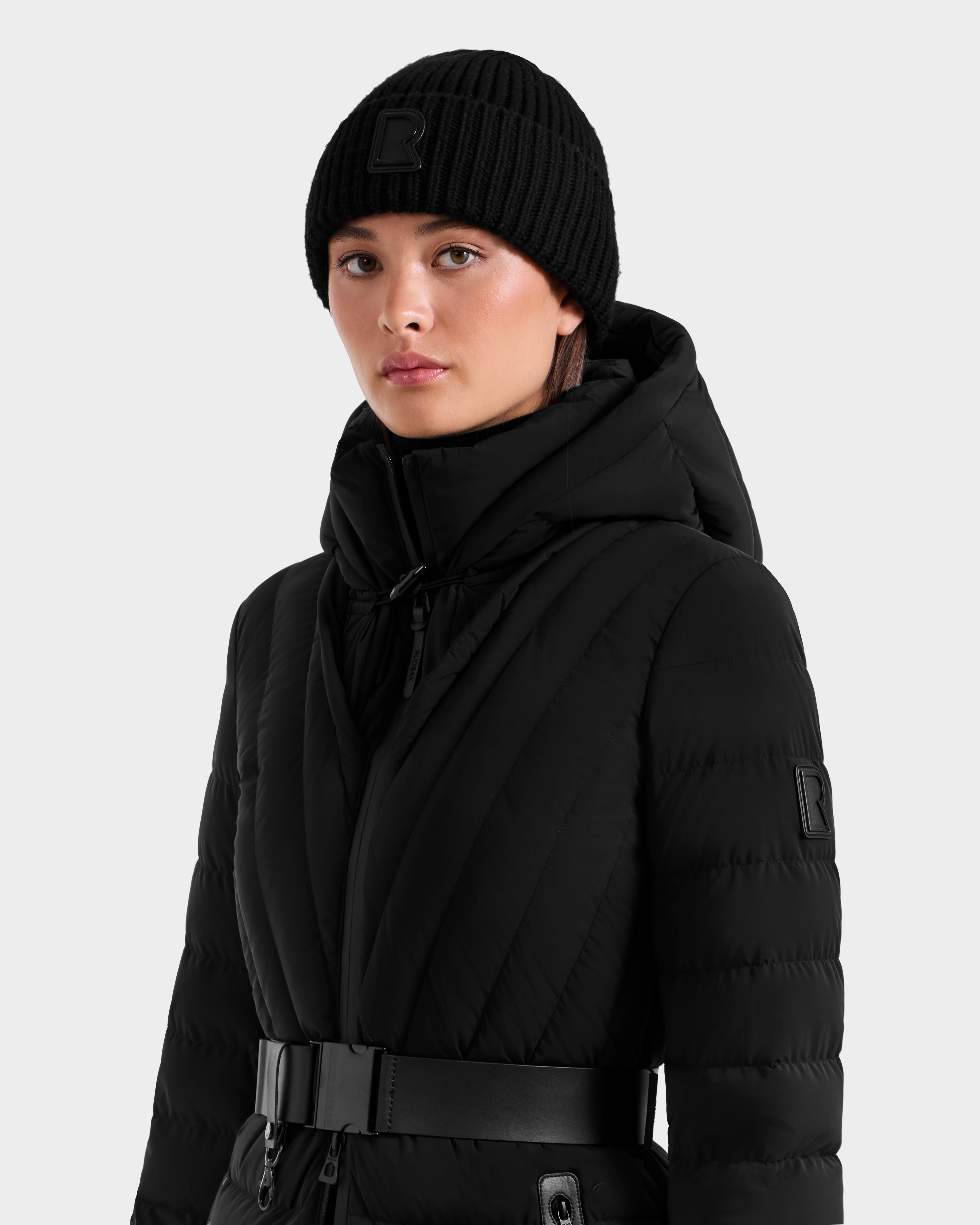 Wearing the Rudsak CYBELLE - 9424504 Black quilted jacket with a high collar, hood, and wide belt, paired with a Rudsak black wool blend beanie, a person poses against a plain light gray background.