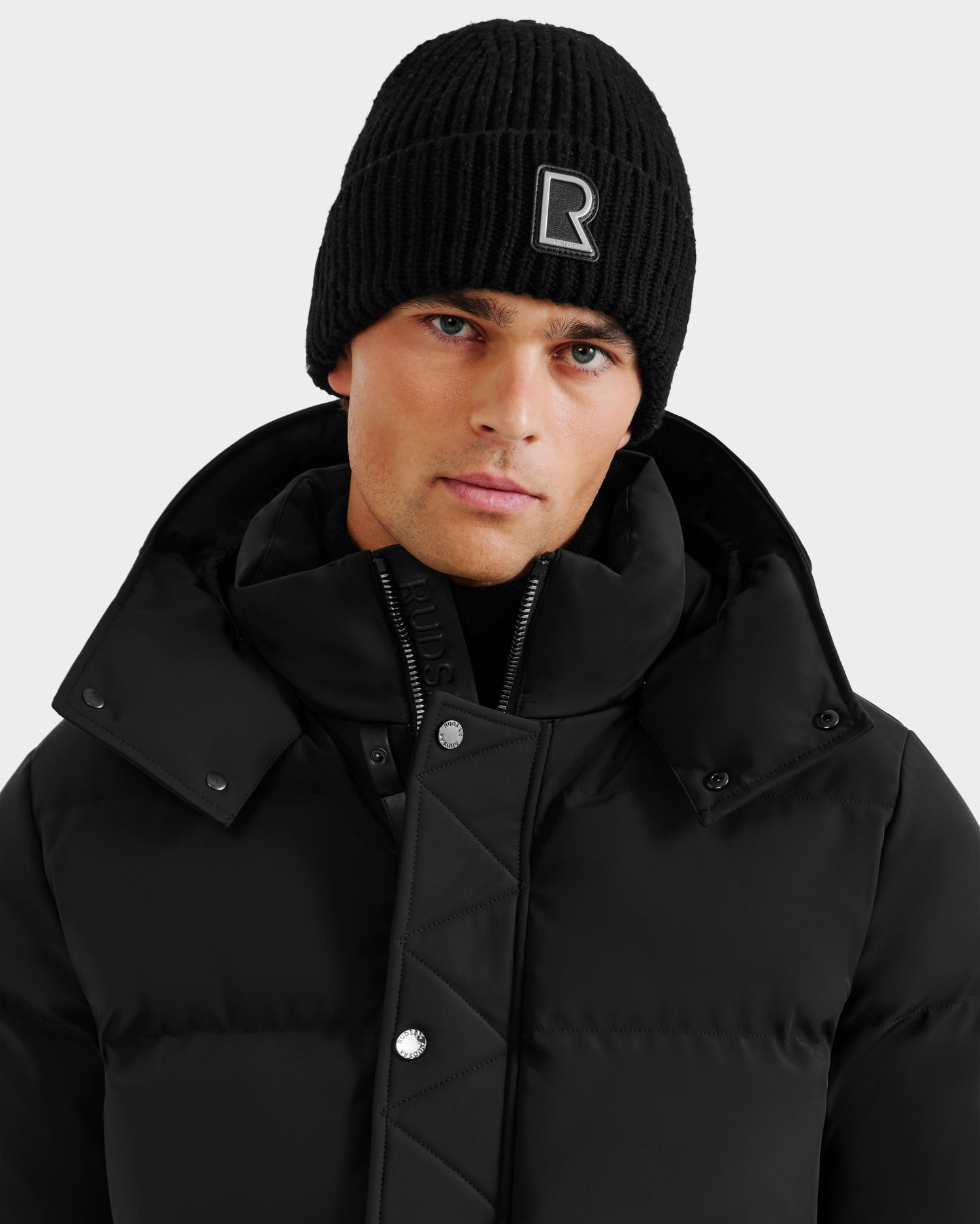 A young man wears the Rudsak CYBELLE - 9424504 Black/Silver quilted winter jacket with a high collar and a knit hat featuring a white R logo, looking directly at the camera against a plain light background.
