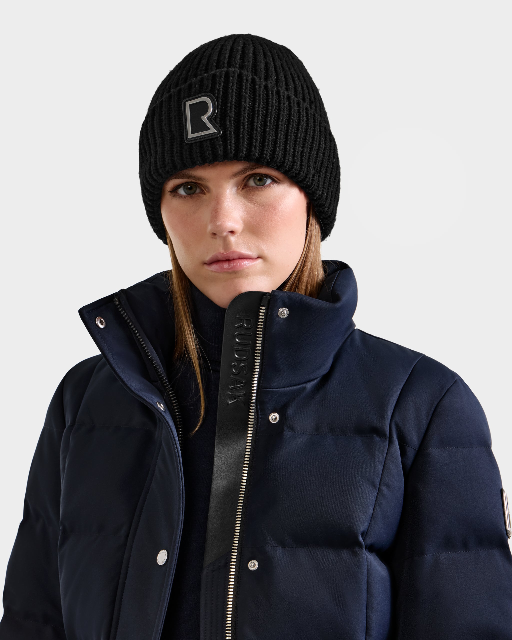 Wearing the Rudsak CYBELLE - 9424504 black wool blend beanie with a bold silver R logo, a woman in a navy puffer jacket stands against a plain light background, looking directly at the camera.