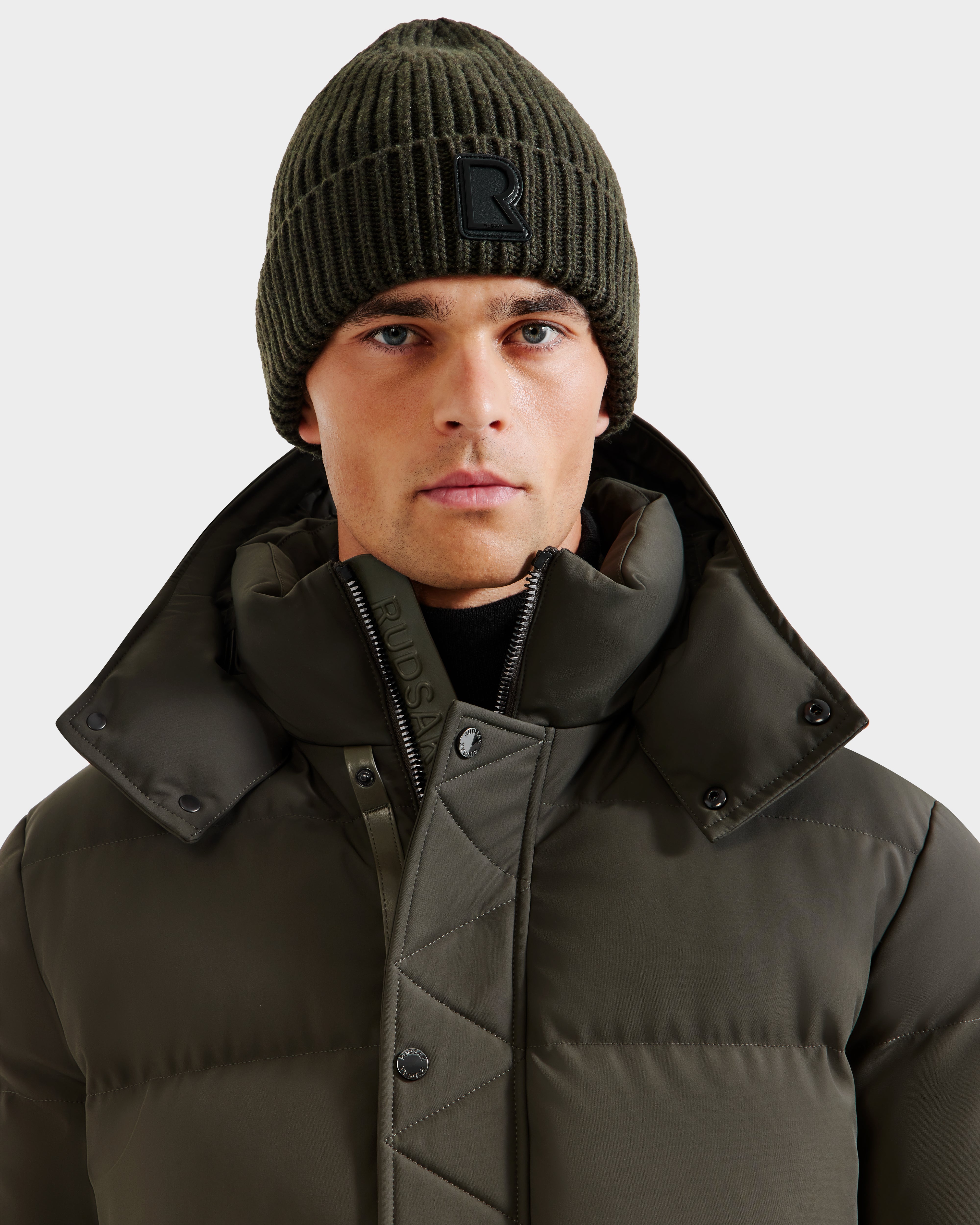 A man wearing the RUDSAK CYBELLE - 9424504 Dark Olive puffer jacket with a high collar and a dark olive ribbed wool beanie poses against a plain light gray background.