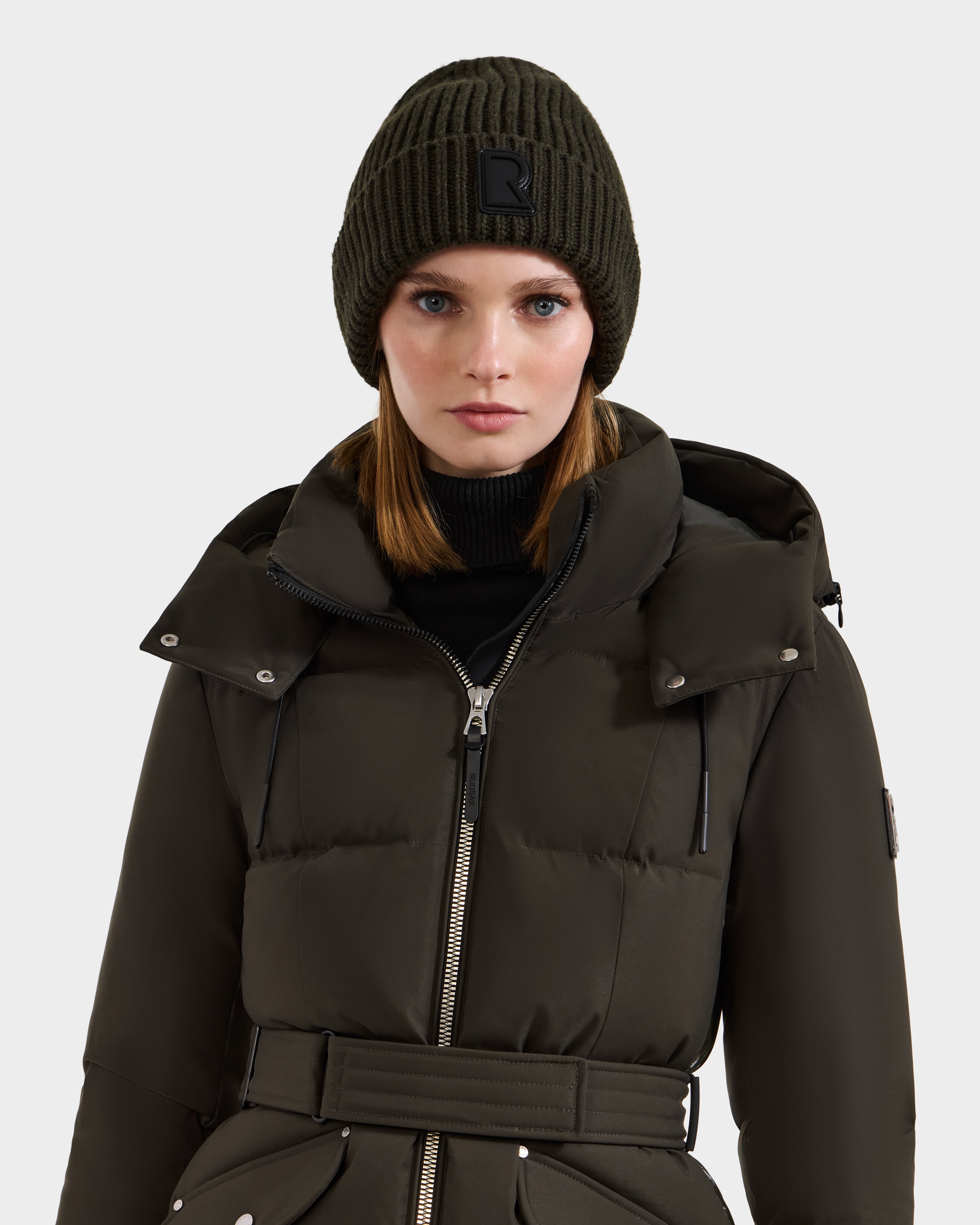A woman models the Rudsak CYBELLE - 9424504 Dark Olive unisex beanie and matching belted puffer coat with hood, standing against a plain light background. Her straight light brown hair and neutral expression complete the winter look.