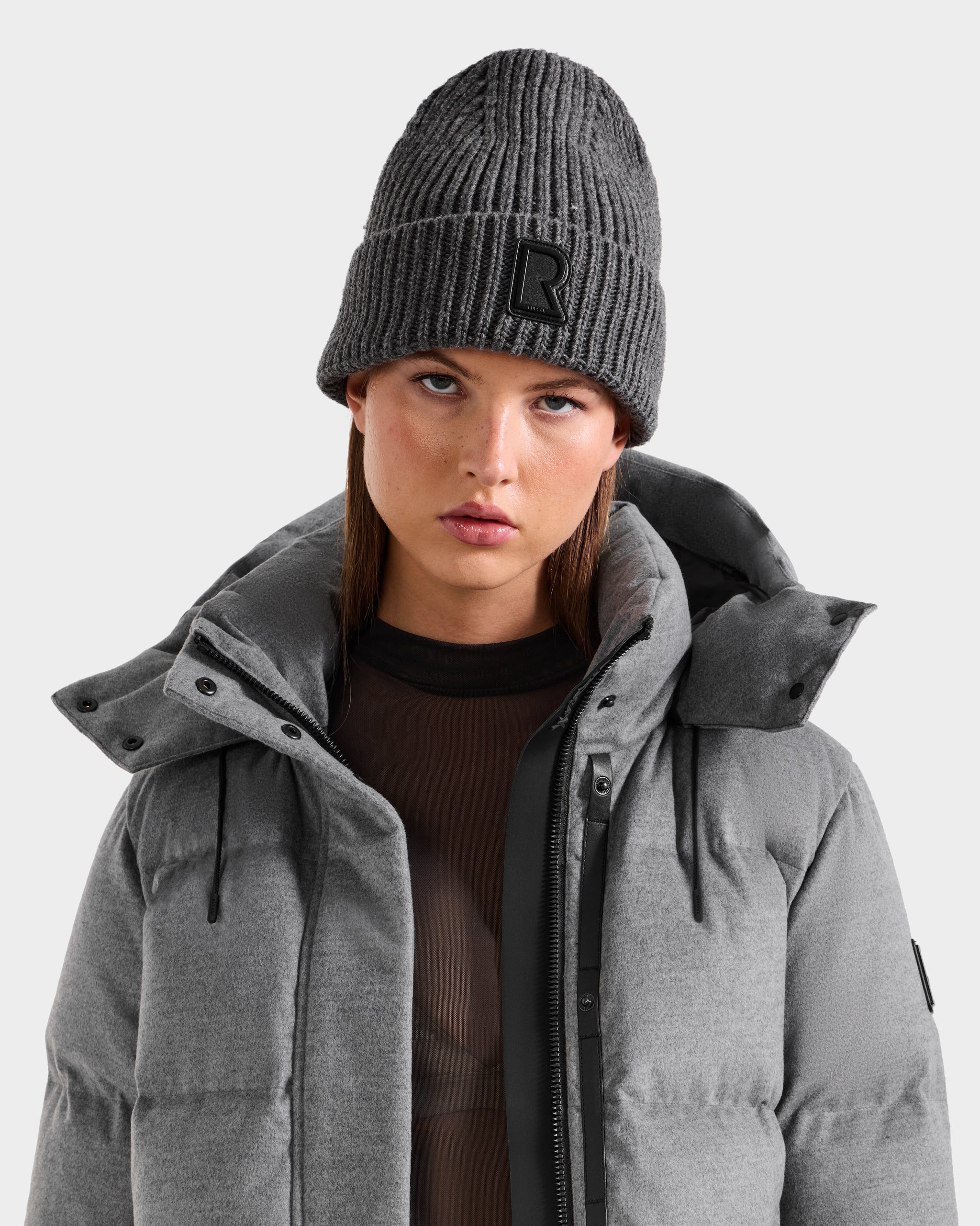 A woman wears the Rudsak CYBELLE - 9424504 Grey puffer jacket with a high collar and a gray wool blend beanie featuring a black “R” patch, looking directly at the camera against a plain light background.