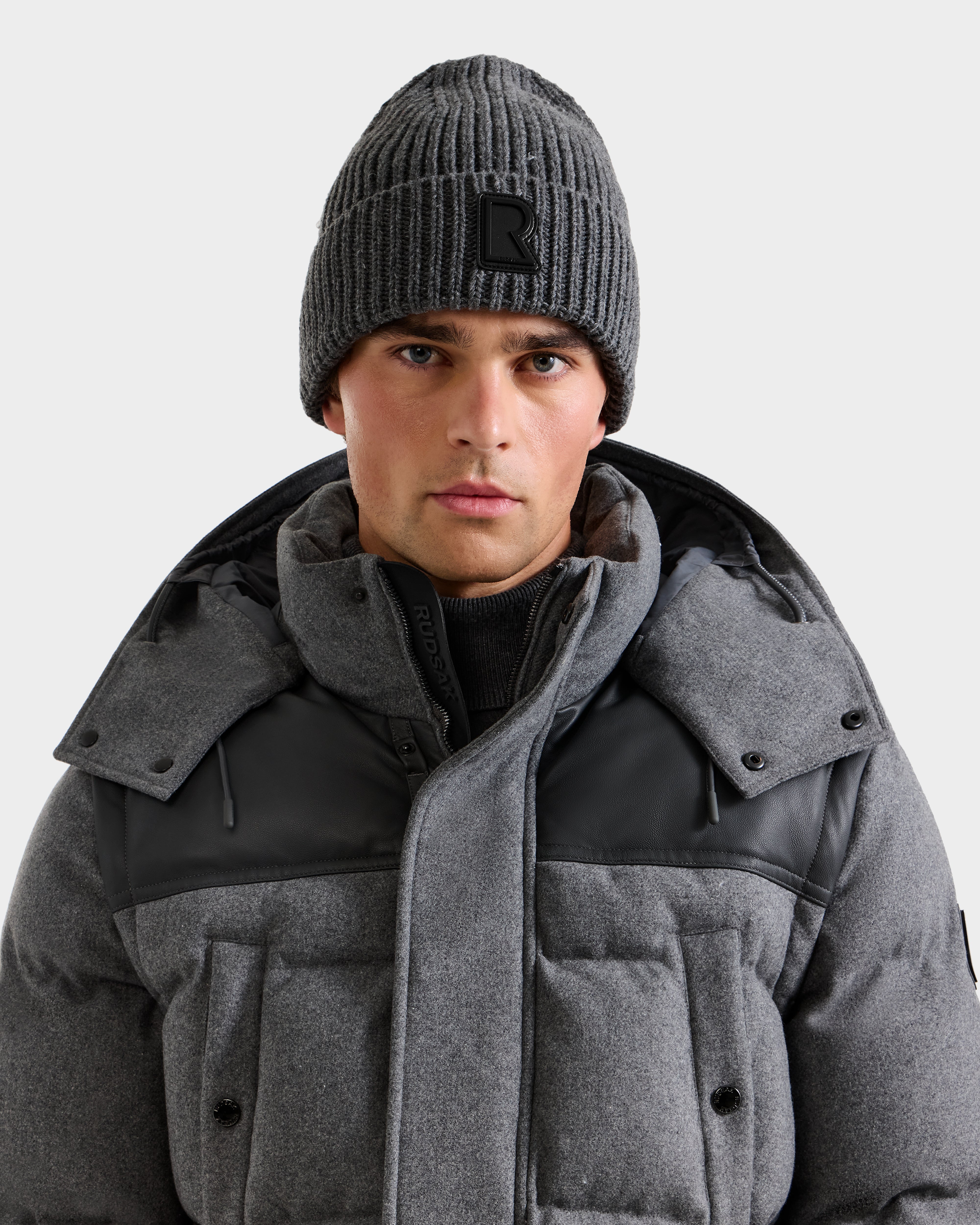 A man faces the camera against a plain light background, wearing the Rudsak CYBELLE - 9424504 Grey padded winter coat with black accents and a luxury knit hat, showcasing stylish Rudsak winter accessories.