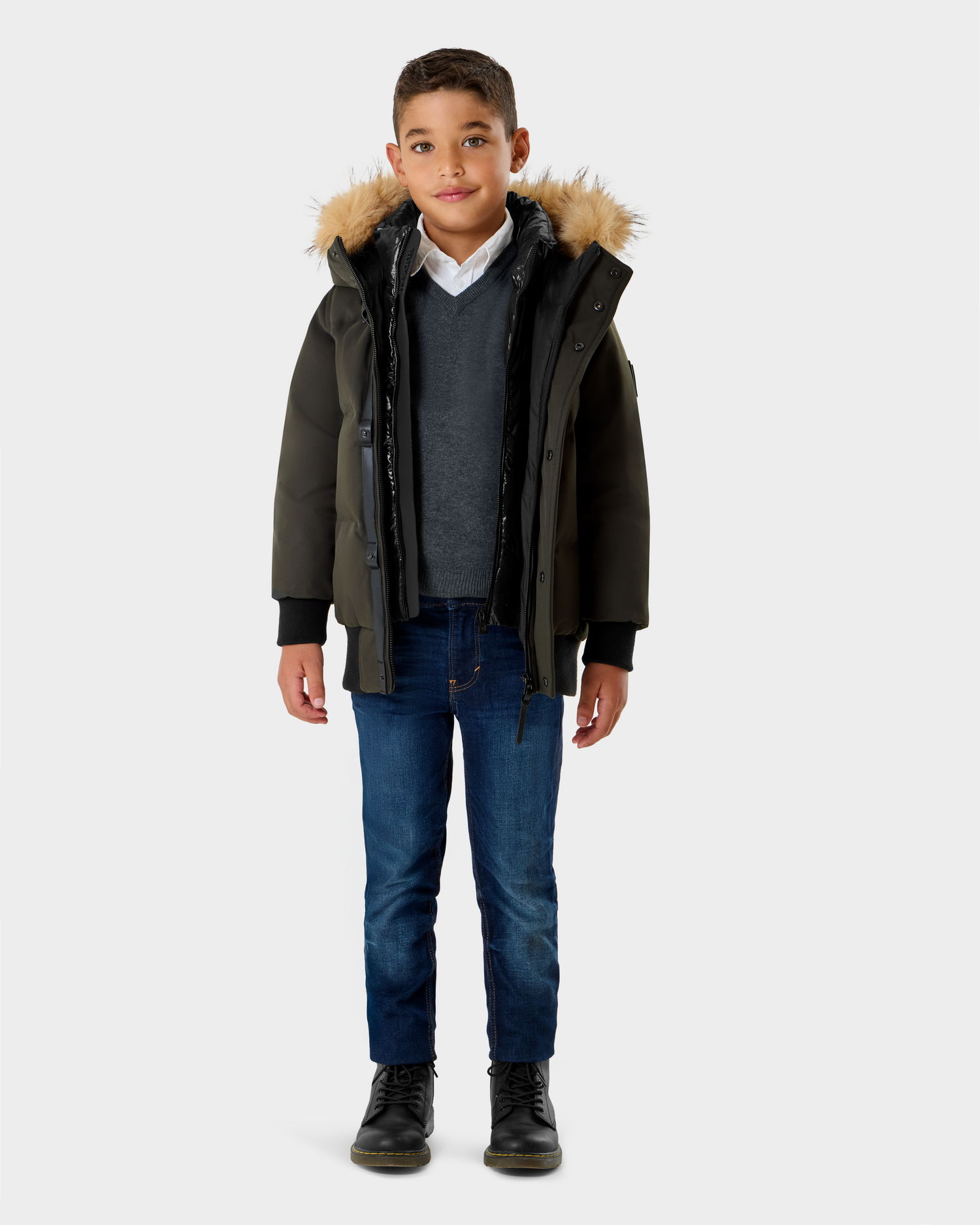 A young boy faces forward in a Rudsak DALLAS K - K125501 Dark Olive parka with a fur-lined hood, gray sweater over white shirt, blue jeans, and black boots against a plain light background.