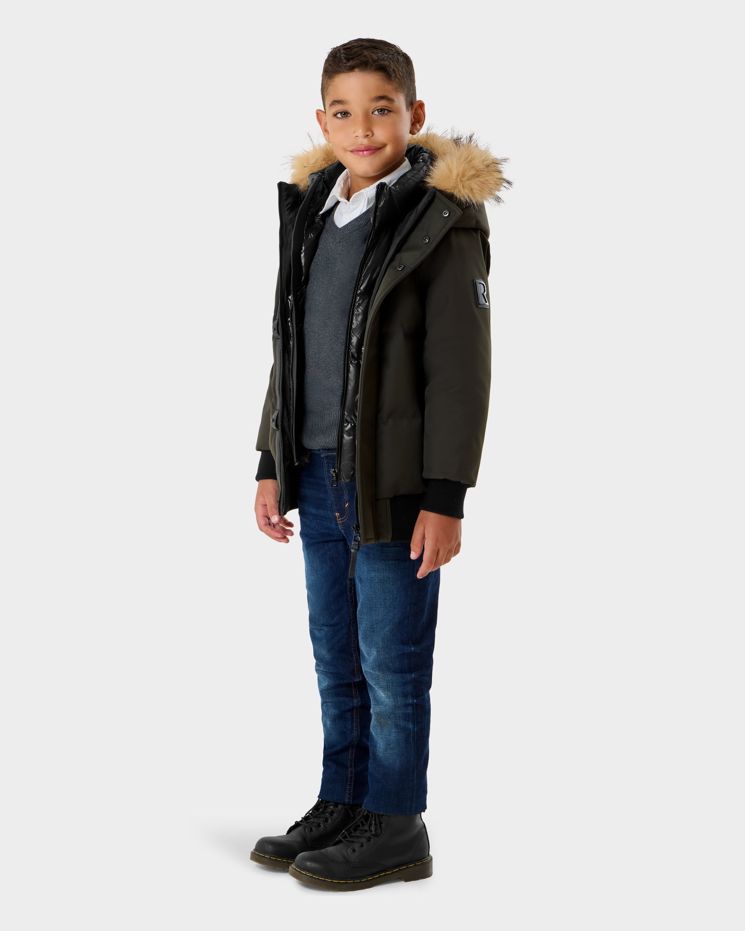 A young boy stands facing forward, wearing the Rudsak DALLAS K - K125501 Dark Olive winter coat with a fur-trimmed hood over a gray sweater, blue jeans, black boots, and a white collared shirt, against a plain light background.