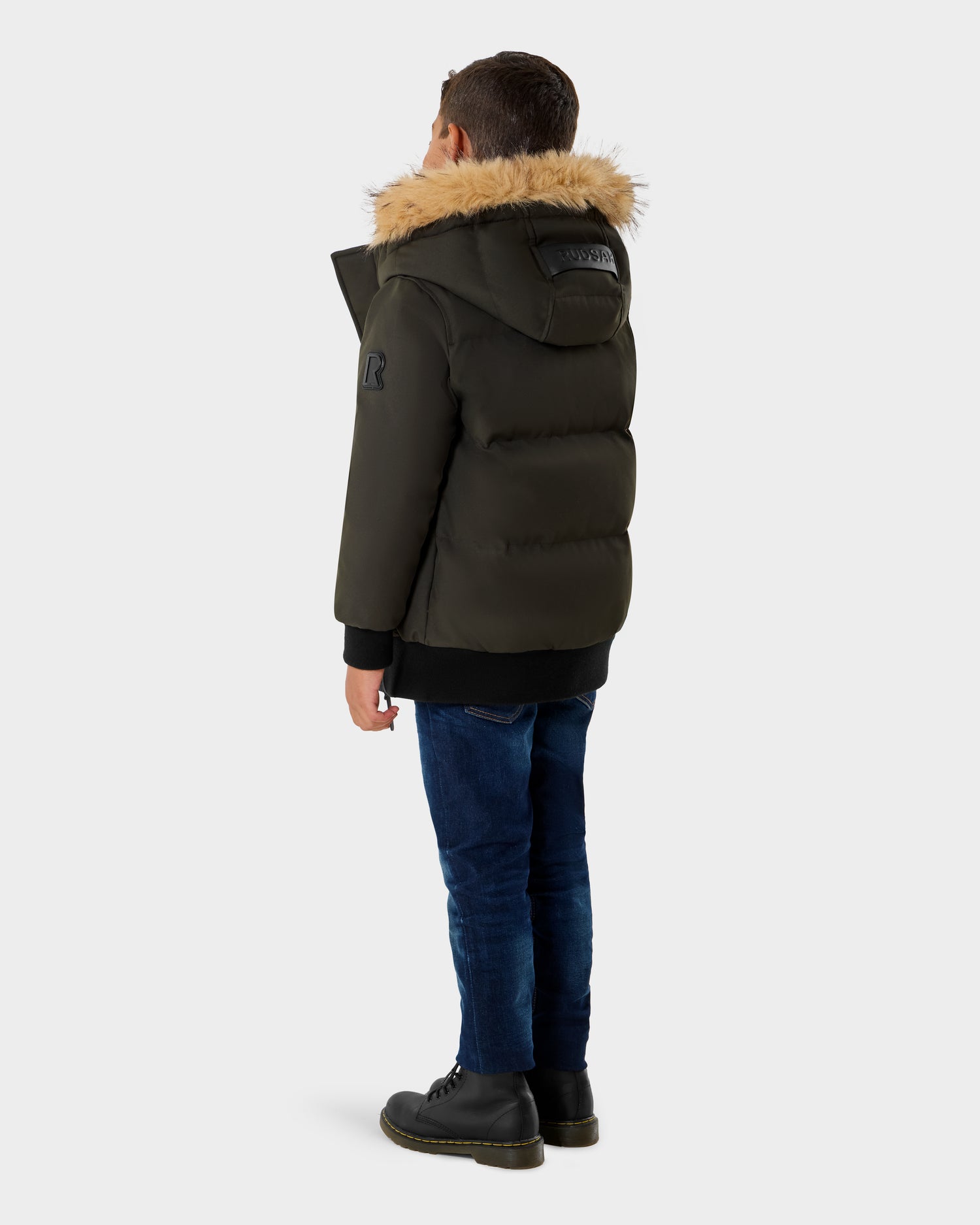 A young boy faces away, wearing the Rudsak DALLAS K - K125501 puffer jacket in dark olive with a faux fur hood, styled with blue jeans and black boots against a plain light background.