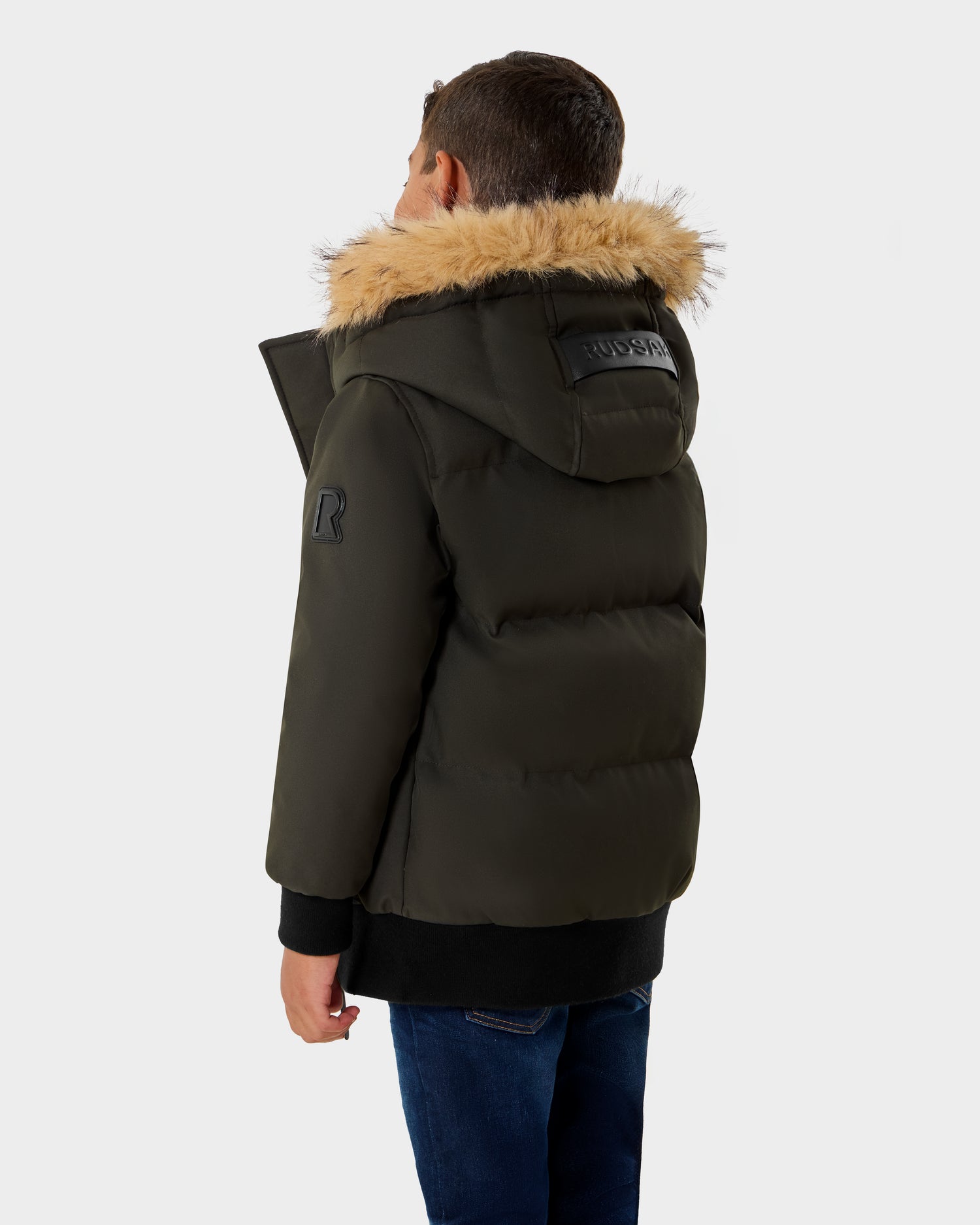 A young boy faces away from the camera, dressed in Rudsak's DALLAS K - K125501 Dark Olive puffer jacket with a faux fur-lined hood and blue jeans, set against a plain, light background.