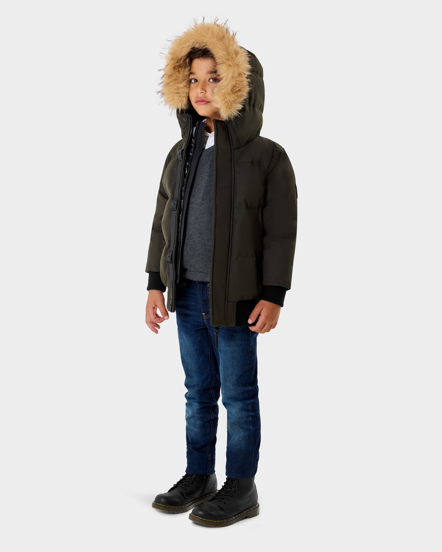 A young boy stands facing forward in a Rudsak DALLAS K - K125501 Dark Olive winter coat with a fur-lined hood, blue jeans, and black boots, looking slightly to the side against a plain light background.