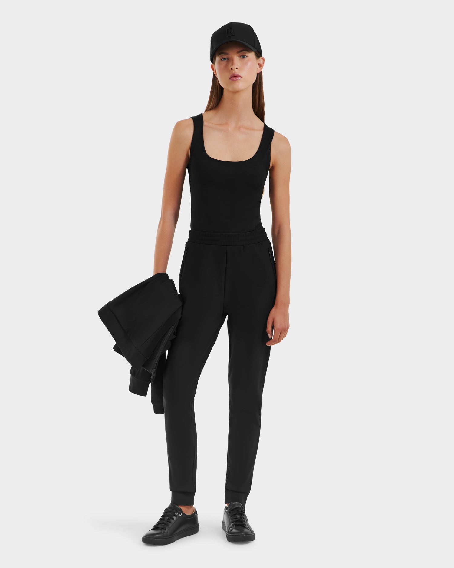 A woman showcases casual athleisure style in the Rudsak DAMERIS - 8525575 Black tank top, paired with joggers, black sneakers, and a cap, holding a black jacket against a plain white background.