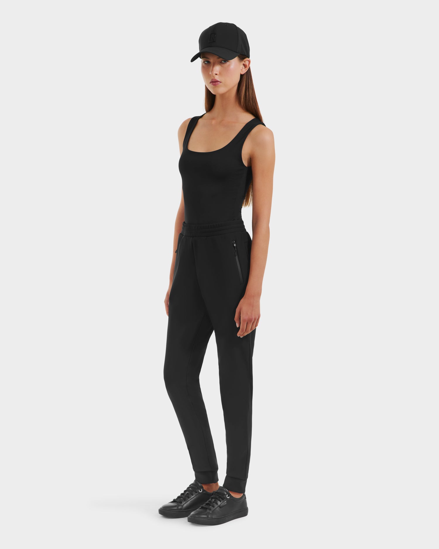 A woman with long straight hair and a neutral expression models the Rudsak DAMERIS - 8525575 Black joggers against a plain background, paired with a black cap, tank top, and sneakers for a sleek athleisure look.