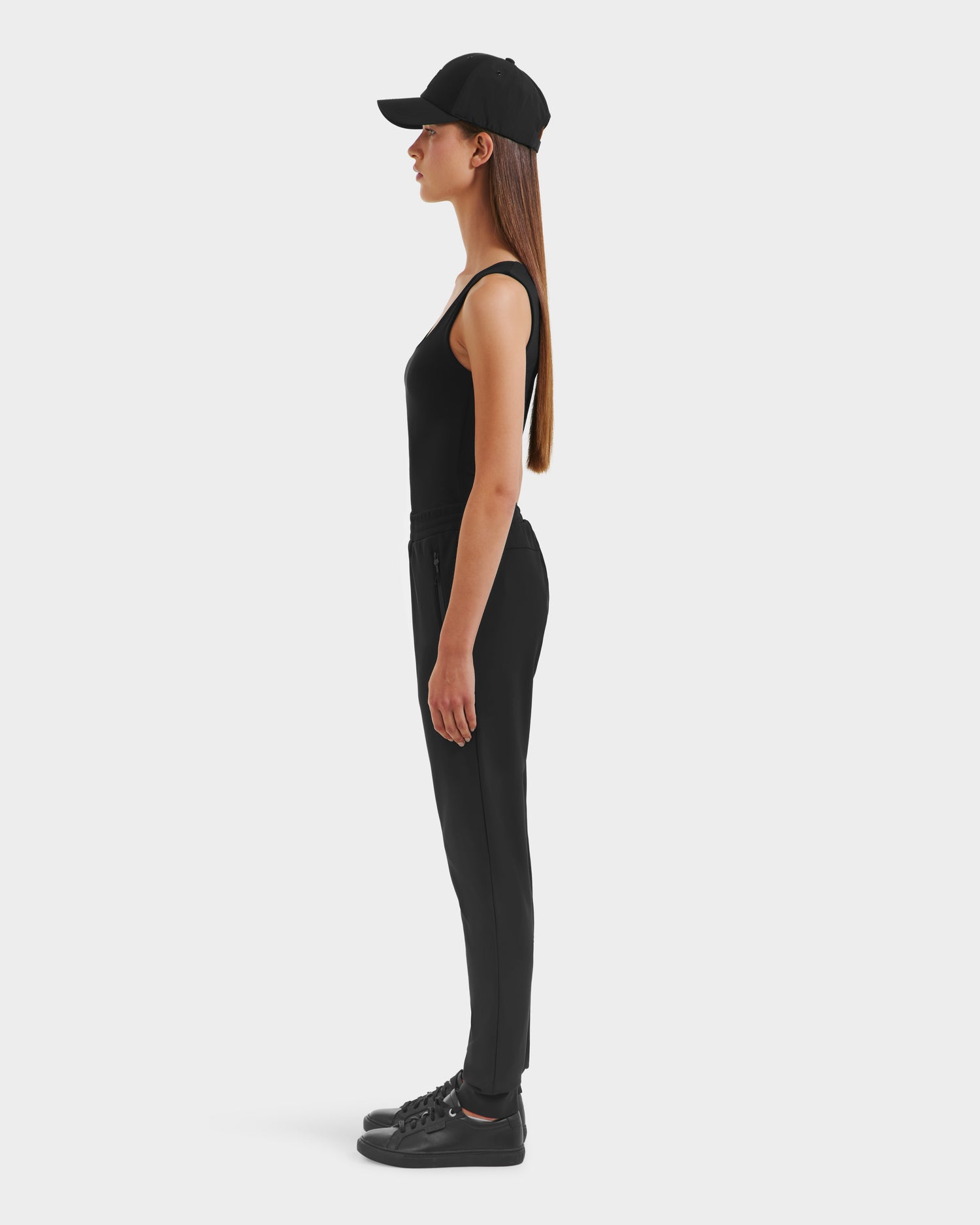 A woman in profile faces left, wearing the Rudsak DAMERIS - 8525575 Black joggers, a sleeveless black top, black cap, and sneakers, styled in athleisure against a plain white background.
