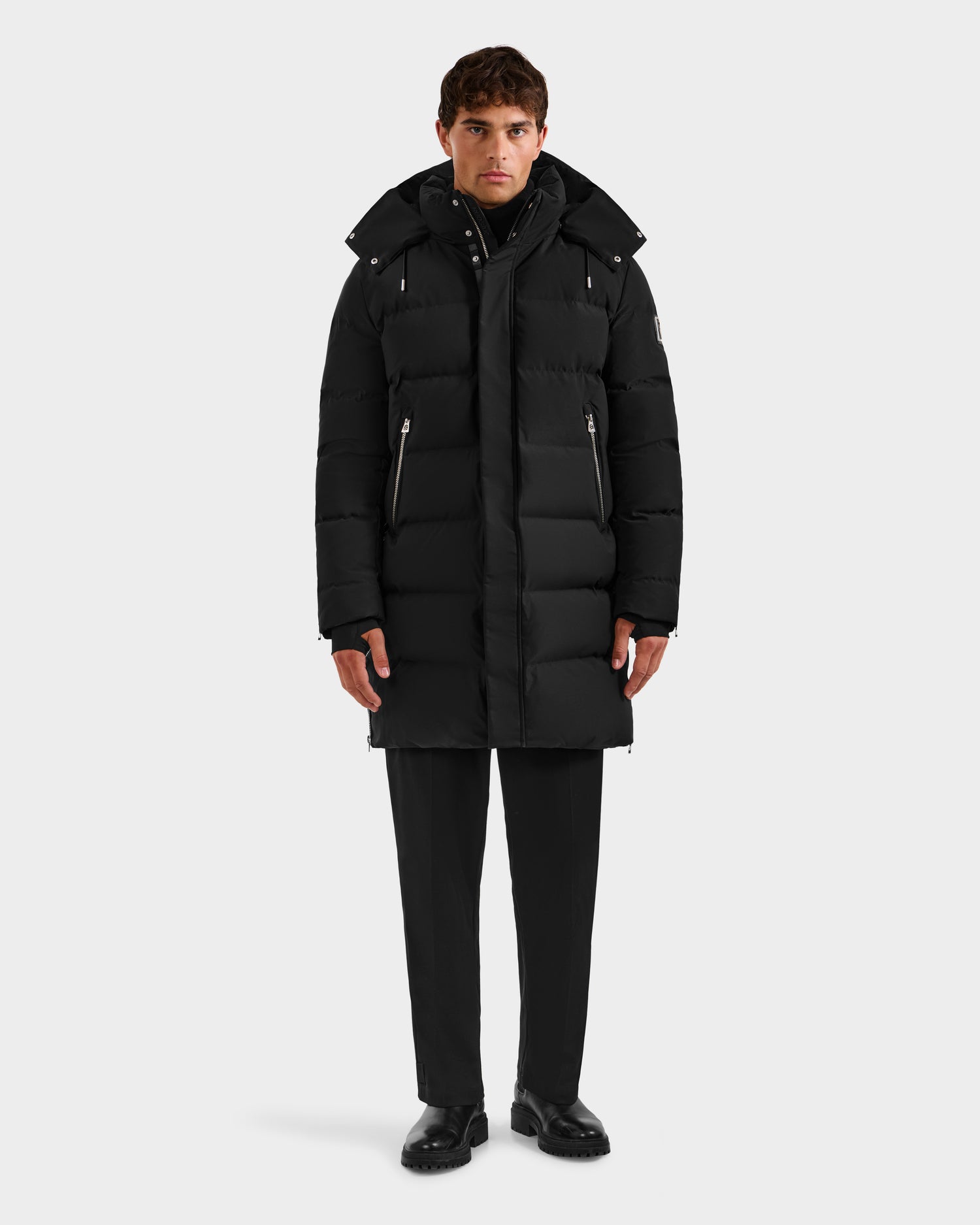 A man stands facing forward in the Rudsak DAO H - 6125583 Black, a long hooded black puffer coat, paired with black pants and shoes, modeling stylish winter outerwear against a plain white background.