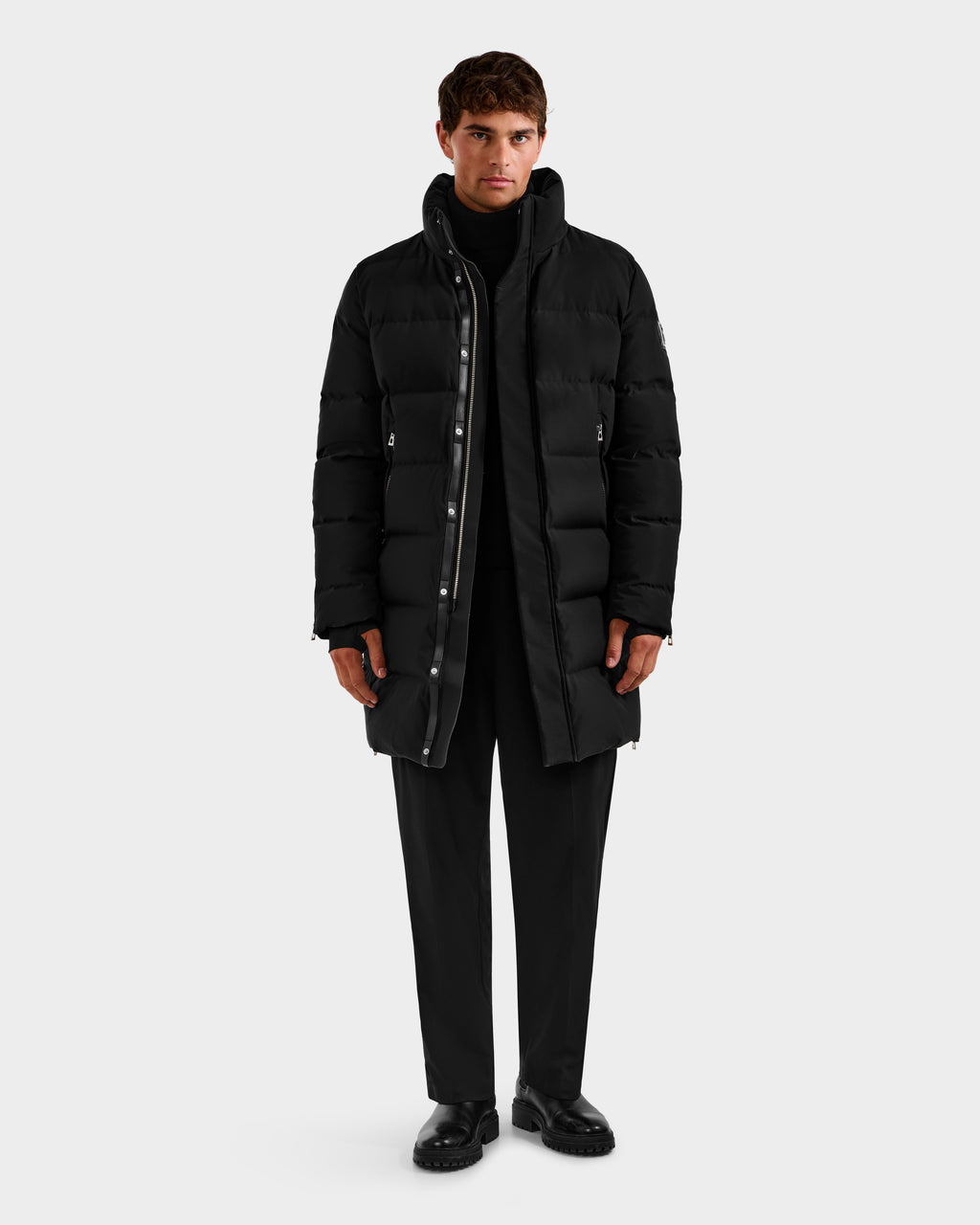 A person faces forward wearing the DAO H - 6125583 Black quilted down jacket by Rudsak over a black turtleneck, pants, and shoes—highlighting sleek winter style against a plain light gray background.