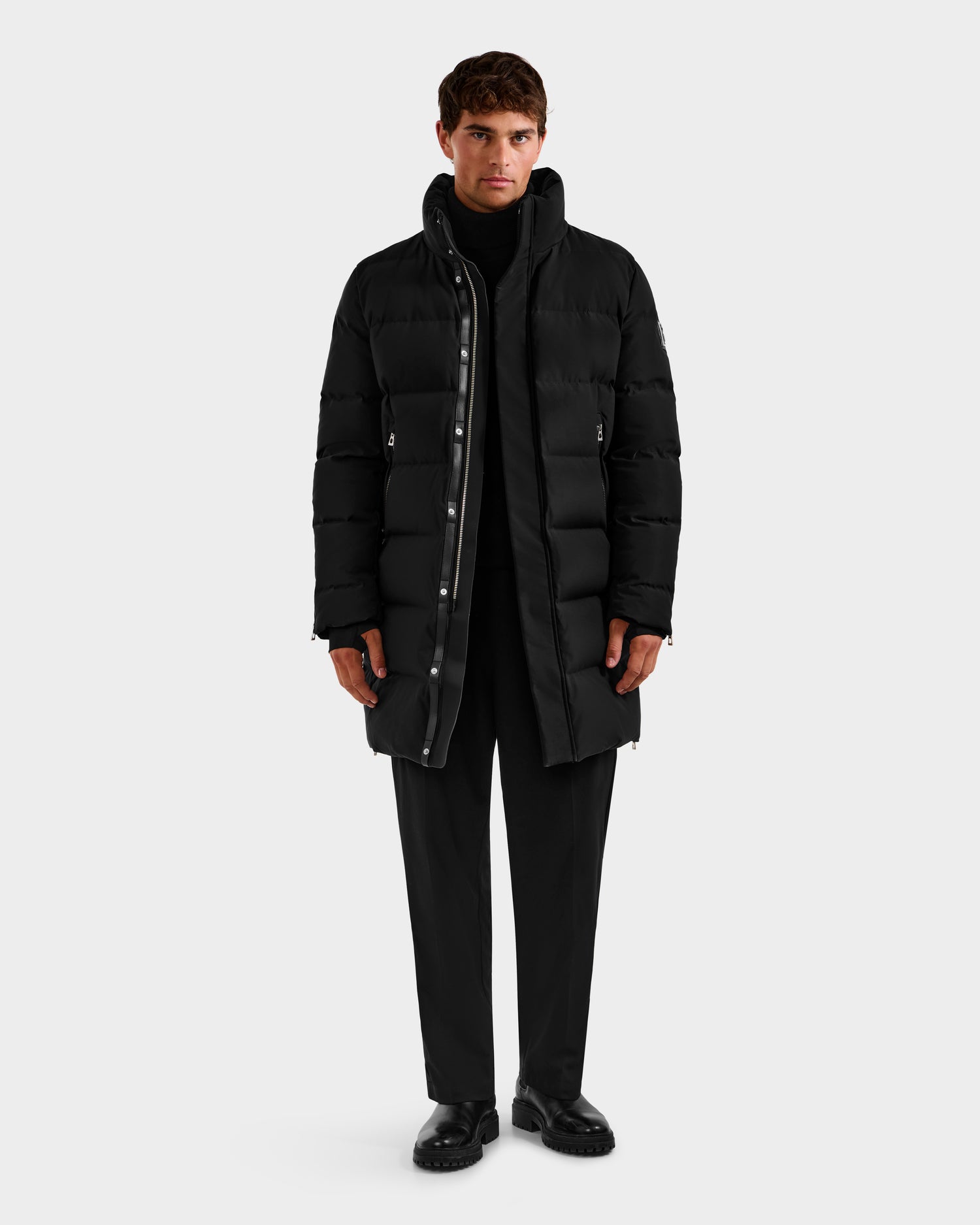 A person faces forward wearing the DAO H - 6125583 Black quilted down jacket by Rudsak over a black turtleneck, pants, and shoes—highlighting sleek winter style against a plain light gray background.