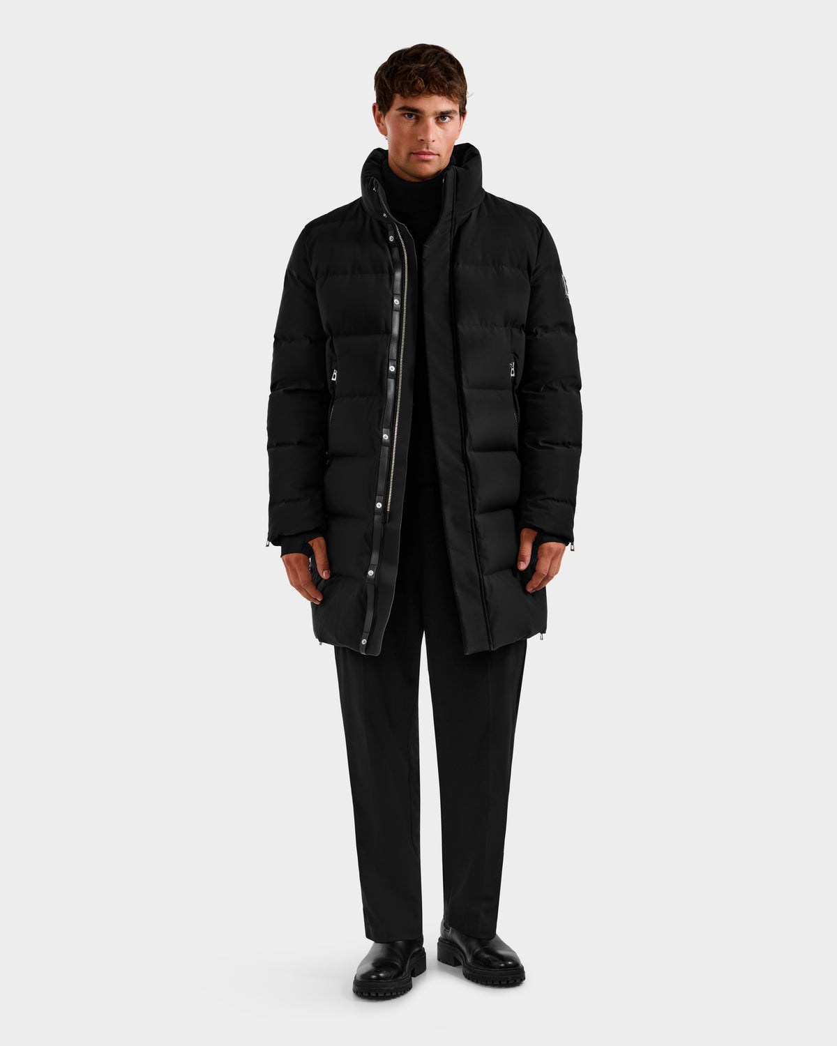 A person faces forward wearing the DAO H - 6125583 Black quilted down jacket by Rudsak over a black turtleneck, pants, and shoes—highlighting sleek winter style against a plain light gray background.