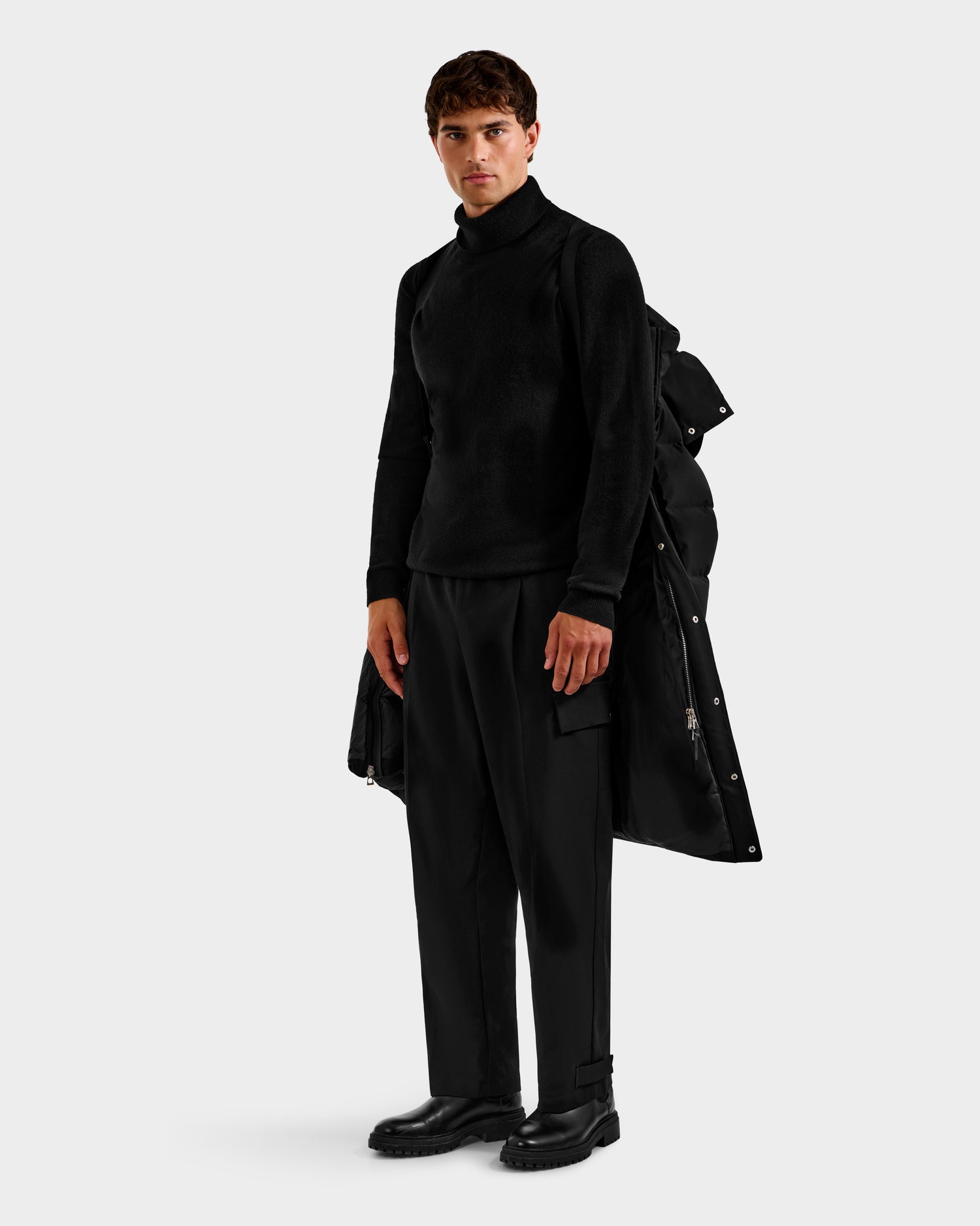 A man wears an all-black outfit featuring the Rudsak DAO H - 6125583 Black winter long coat draped over one shoulder, paired with a turtleneck sweater, loose trousers, and boots against a plain light background.