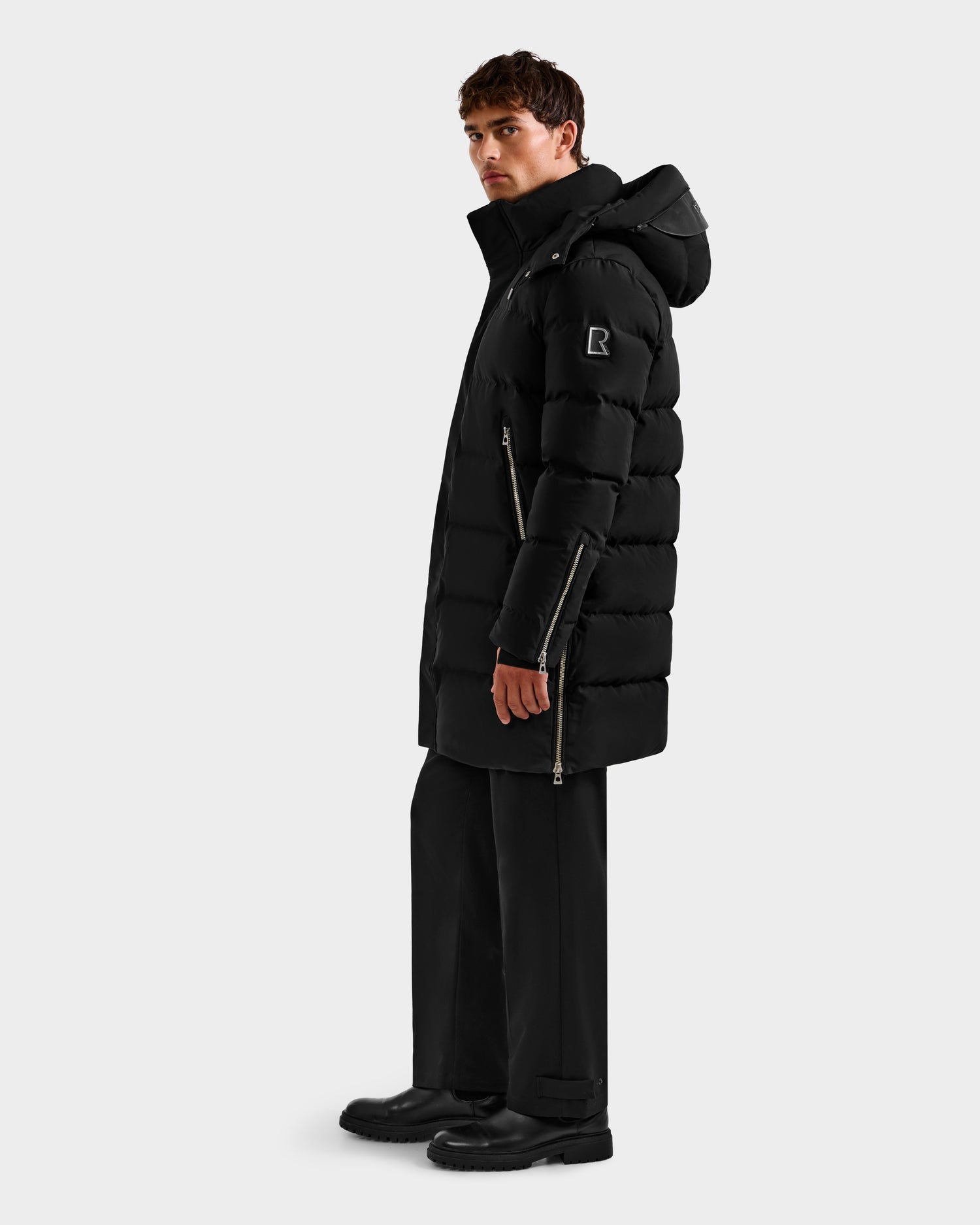 A man stands sideways against a plain light background wearing the Rudsak DAO H - 6125583 Black puffer coat with hood, visible zippers, sleeve patch, black pants, and boots—perfect for cold-weather performance.
