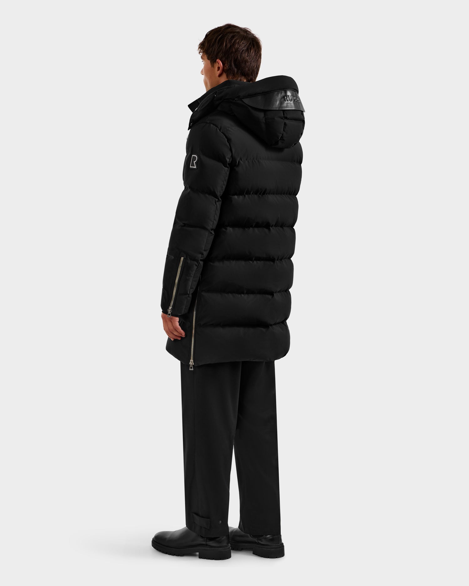 A person faces away, modeling the Rudsak DAO H - 6125583 Black long hooded puffer coat with black pants and boots, set against a plain light gray background.
