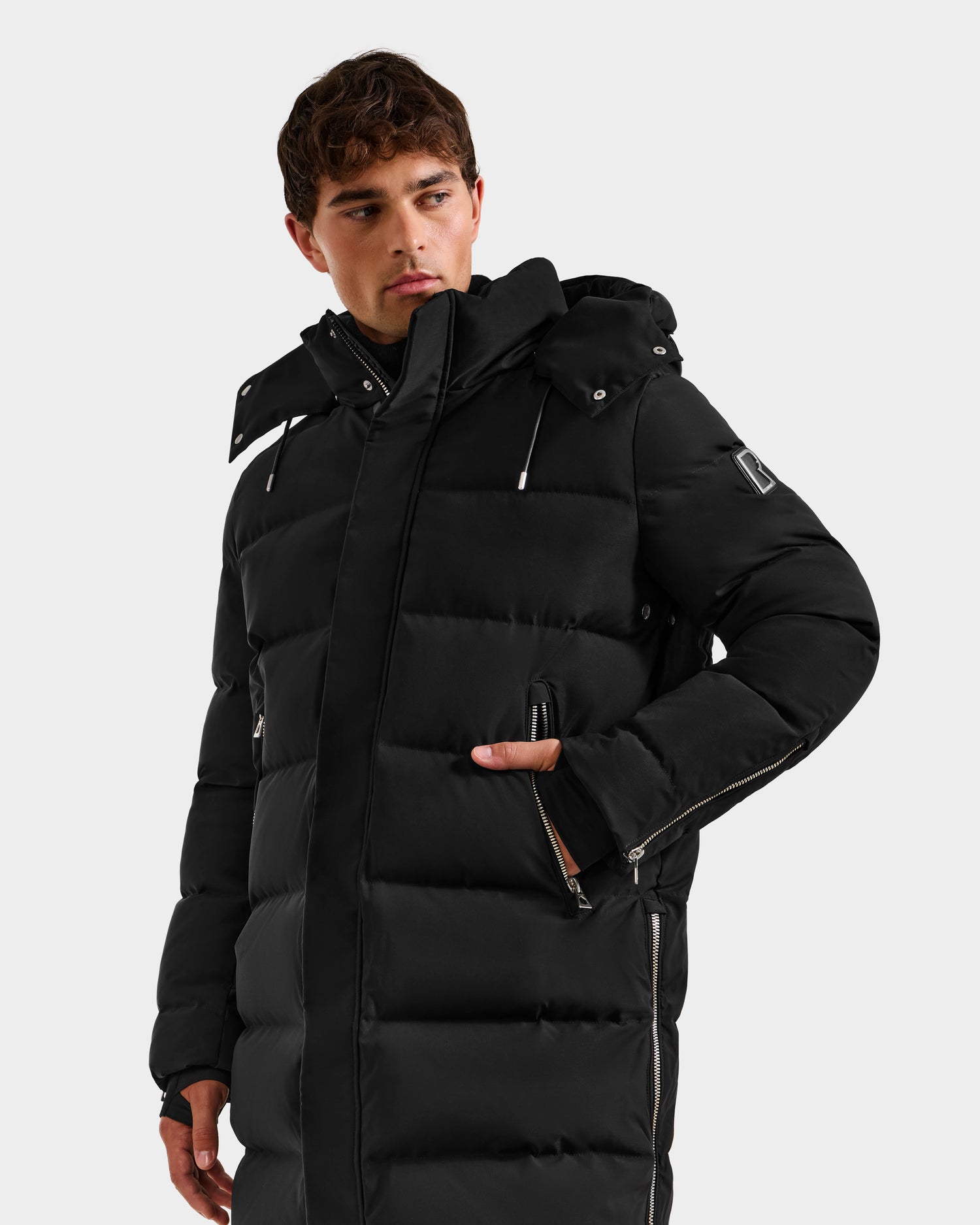 A person wears the DAO H - 6125583 Black men’s quilted down jacket by Rudsak with a hood, against a plain light background, hand in pocket—an ideal winter outerwear choice for cold-weather performance.