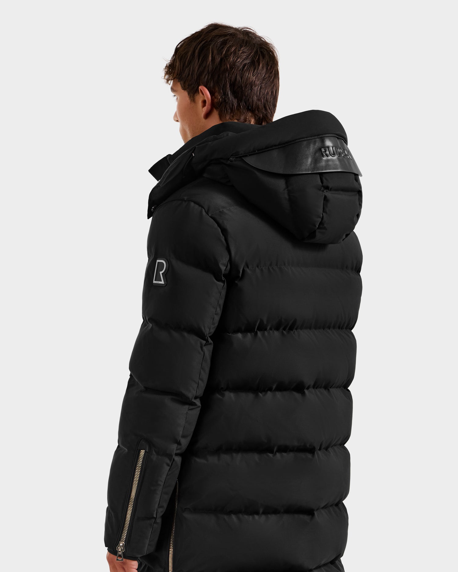 A person wearing the Rudsak DAO H - 6125583 Black men’s quilted down jacket with hood, seen from behind on a white background. Winter outerwear features a logo on the sleeve and zippered cuffs for cold-weather performance.