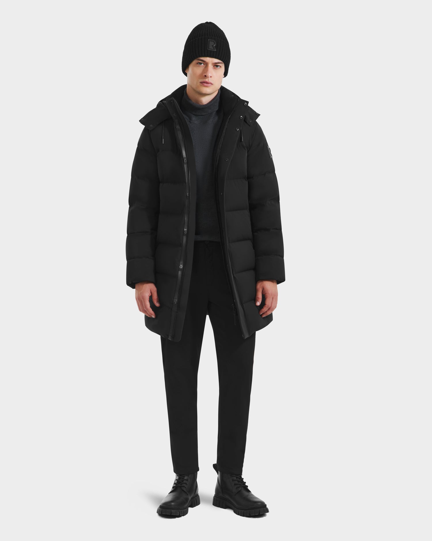 A person stands against a plain background wearing the Rudsak DAO - 6124604 Black mens mid-length puffer, styled with a beanie, turtleneck, pants, and boots. Their arms are relaxed at their sides and their expression is neutral.