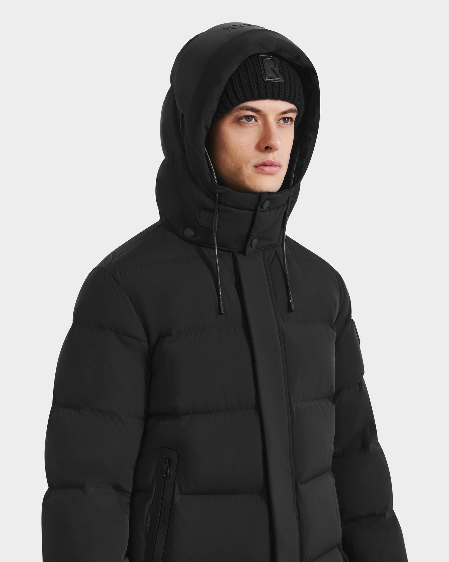 A person wears the Rudsak DAO - 6124604 Black mens mid-length puffer jacket with a weather-resistant hooded shell and a black knit beanie, looking slightly to the side against a light gray background.
