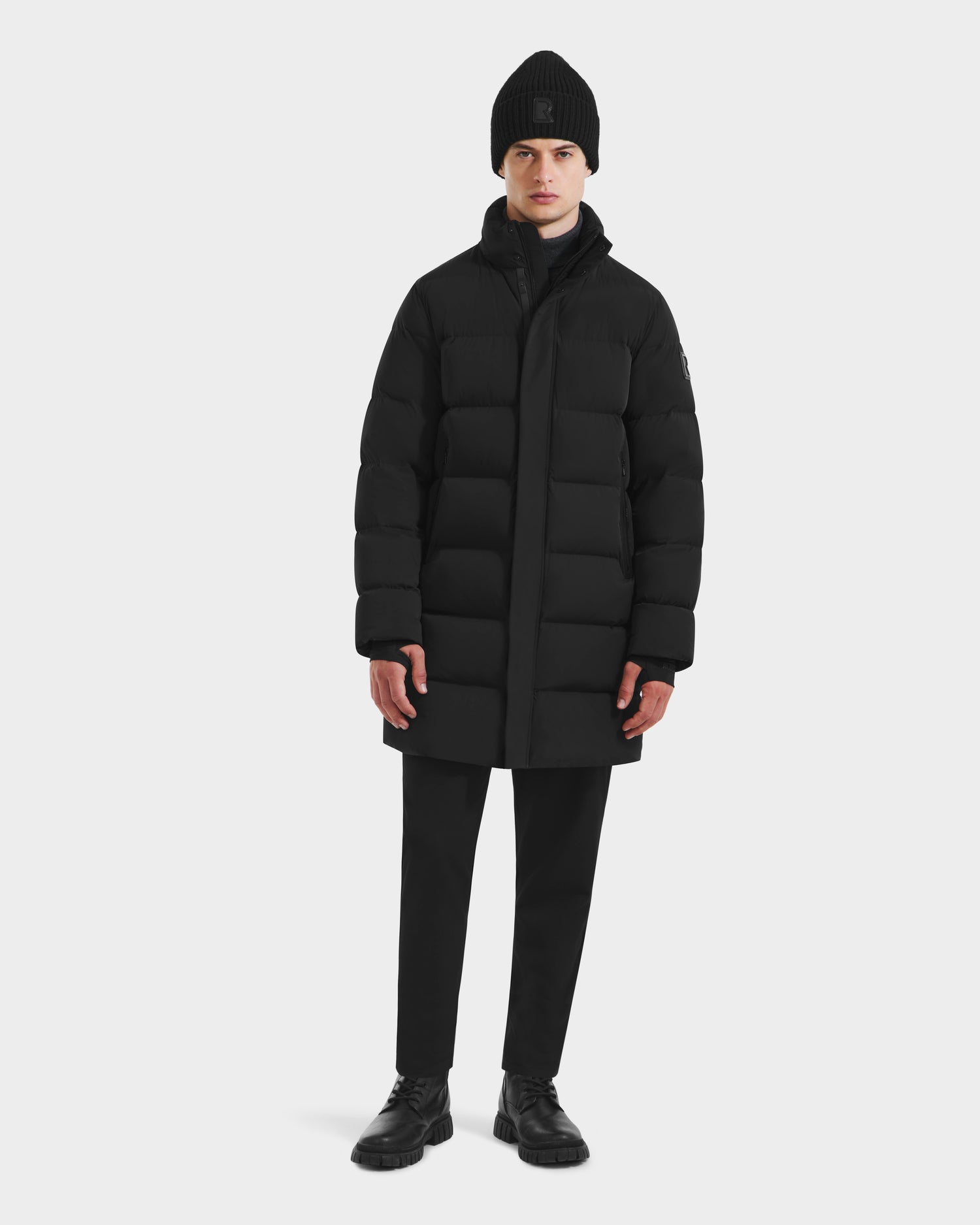 A person stands against a plain background wearing the Rudsak DAO - 6124604 Black mens mid-length down-filled puffer, paired with black pants, a black knit beanie, and black shoes, looking straight ahead with arms relaxed at their sides.