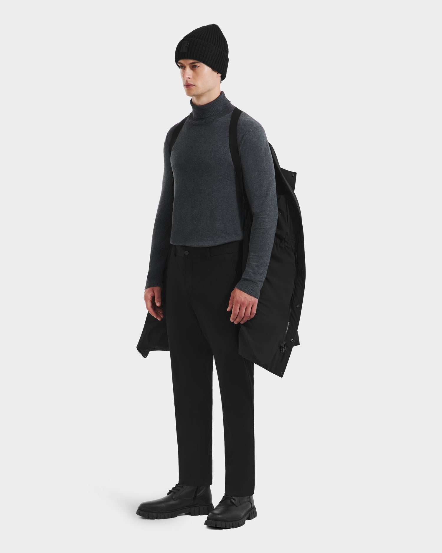 A person stands against a white background with the Rudsak DAO - 6124604 Black mens weather-resistant shell mid-length puffer draped over one shoulder, wearing a black beanie, dark gray turtleneck, black pants, and black shoes.