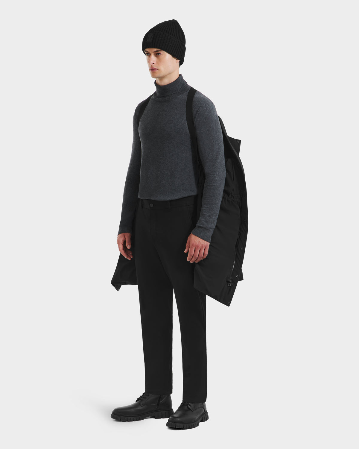 A person stands against a white background with the Rudsak DAO - 6124604 Black mens weather-resistant shell mid-length puffer draped over one shoulder, wearing a black beanie, dark gray turtleneck, black pants, and black shoes.