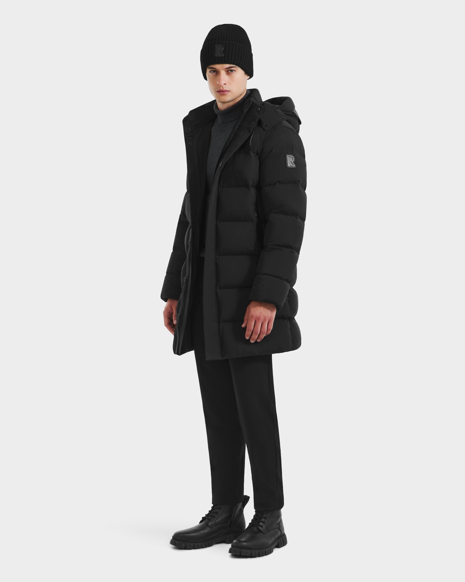 A man wearing the Rudsak DAO - 6124604 Black mid-length puffer stands against a plain white background, styled with a black beanie, black pants, and black boots, looking slightly to the side.