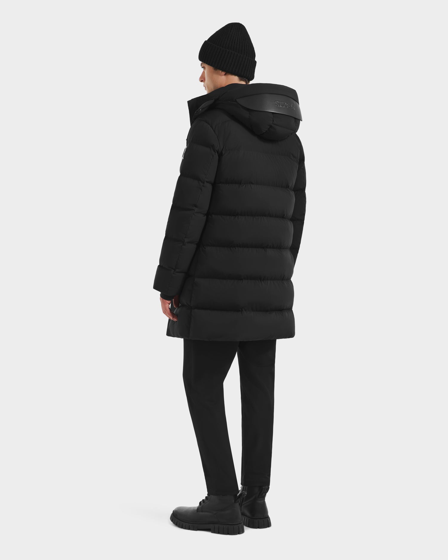 A person wears the Rudsak DAO - 6124604 Black men’s mid-length puffer with a weather-resistant shell, plus a black beanie, pants, and shoes, standing sideways against a plain white background.