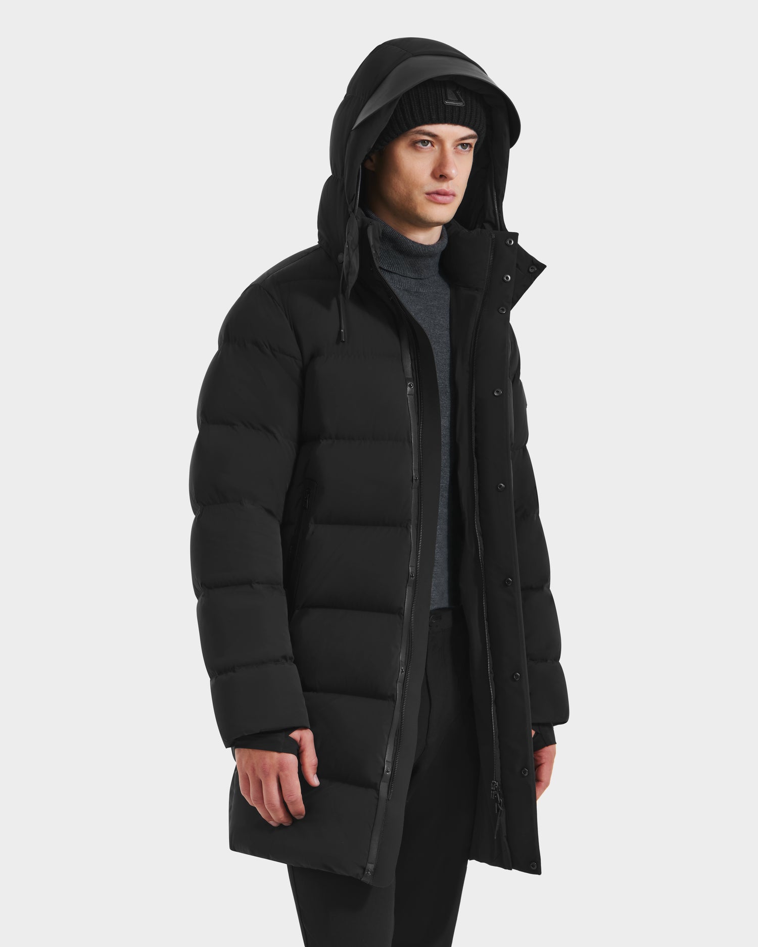 A person wearing the Rudsak DAO - 6124604 Black men’s puffer jacket with a weather-resistant shell and hood over a dark turtleneck and knit hat, standing against a plain white background.